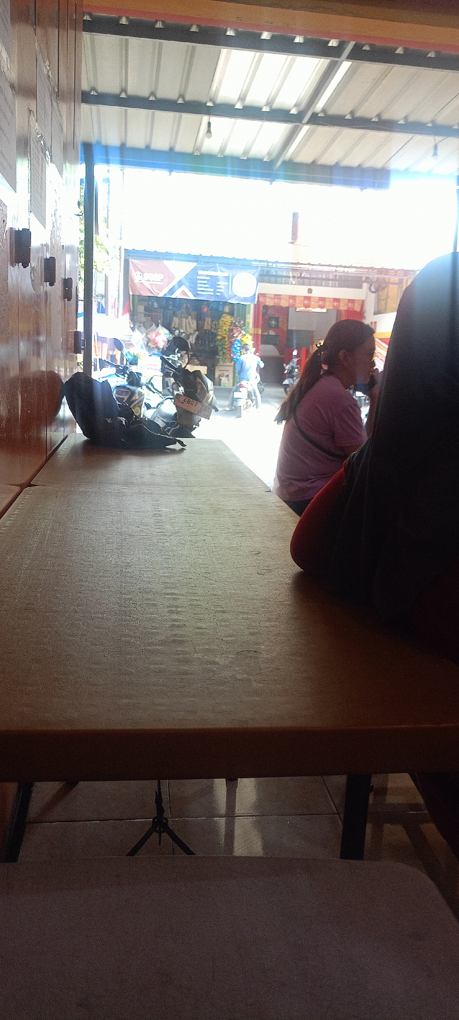 A woman with her back to the camera is sitting at a table outdoors in a bustling street market setting. She is wearing a light purple t-shirt and her hair is pulled back into a ponytail. To her left, a motorcycle is parked, and further down the street, other motorcycles and people are visible, suggesting a busy commercial area. The scene is lit by bright daylight filtering through a corrugated metal awning. In the foreground, a plain wooden table occupies most of the frame, with a shadow of a tripod visible underneath. The overall impression is a snapshot of everyday life in a vibrant Indonesian street market.