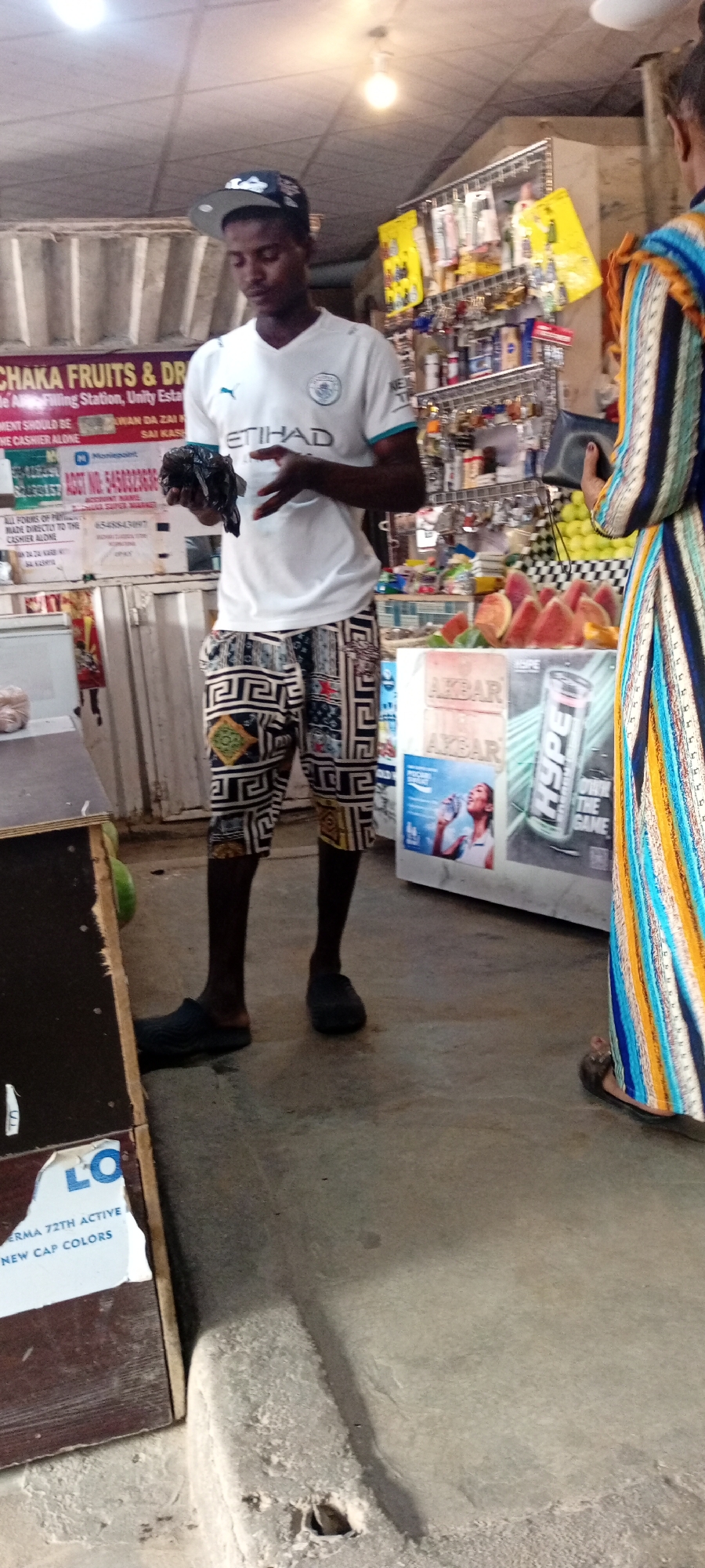 In a dimly lit convenience store in Aviation Quarters, Nigeria, a young man stands holding a dark plastic bag. He is wearing a white Manchester City football jersey with the "Etihad" sponsor and a baseball cap. His shorts feature a distinctive geometric pattern. To his right, another person, partially visible and wearing a vibrantly striped dress, is also in the store. Behind them, shelves are stocked with various items, including drinks, snacks, and possibly toiletries. Sliced watermelons are displayed on a counter, and a tall can with "Hype" and "Own The Game" branding is visible. A sign on a wooden surface reads "CHAKA FRUITS & DR" and mentions "Wing Station, Unity Esta," along with instructions about payments and a "SUPER MARKET" account. Another sign indicates "ERMA 72TH ACTIVE NEW CAP COLORS." The overall impression is of a typical day in a local Nigerian convenience store.