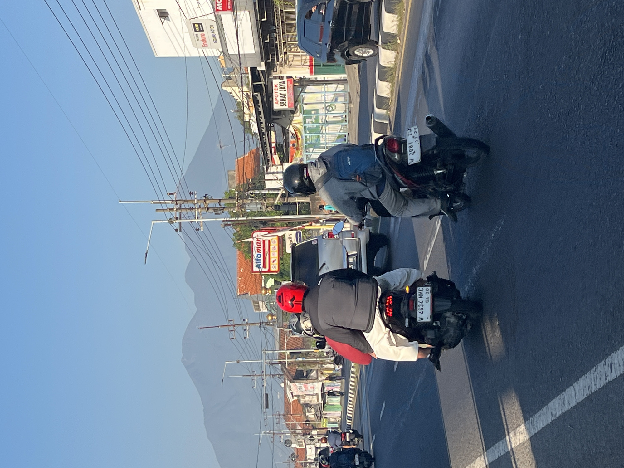 The image depicts a street scene in Siring, Indonesia, with a clear blue sky and mountains in the background. Two motorcycles are prominently featured in the foreground, traveling on a paved road. The first motorcycle, on the left, has two people riding it. The rider wears a black jacket and a red helmet with a mustache design. The passenger is wearing white pants and a red top. The license plate is visible as W 4634 NHC. The second motorcycle, on the right, also has two individuals. The rider wears a gray hooded jacket and a black helmet, with a blue backpack. The passenger is not fully visible. This motorcycle's license plate reads W 2081 ZW.
Several buildings line the right side of the street, including a white building with signage for "Enduro" and "MC" (Motorcycle). Another building displays "Alfamart" and "APOTEK SEHAT JAYA". Power lines and utility poles are visible overhead. A silver van is parked on the right side of the road, and a black SUV is also present further down. Other vehicles and individuals on motorcycles are observed in the middle ground, suggesting moderate traffic. The road is marked with white lines, and a concrete barrier with black and white markings separates the traffic lanes. The environment appears to be a commercial or urban area.