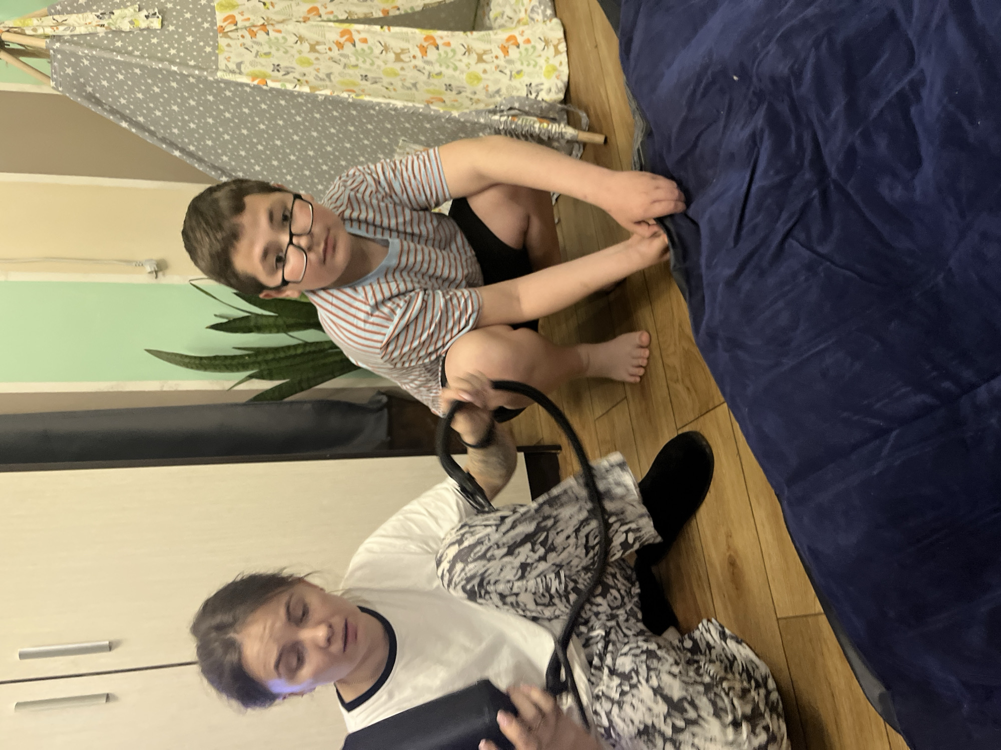 Location: Oryol-Izumrud, Russia.
A young boy wearing glasses and a striped shirt is kneeling on a wooden floor, near a teepee-style structure with patterned fabric. He is facing the camera and appears to be reaching towards a large, dark blue inflatable mattress. To the left of the boy, a woman wearing a white shirt with a black collar, black leggings, and what appears to be a black and white patterned dress or skirt is sitting and looking down, possibly holding a device in her hands. She also has a black object, possibly a headphone or microphone, in her lap. The background includes a cupboard or drawers and a snake plant, the floor is wooden, and the tent is partially visible.