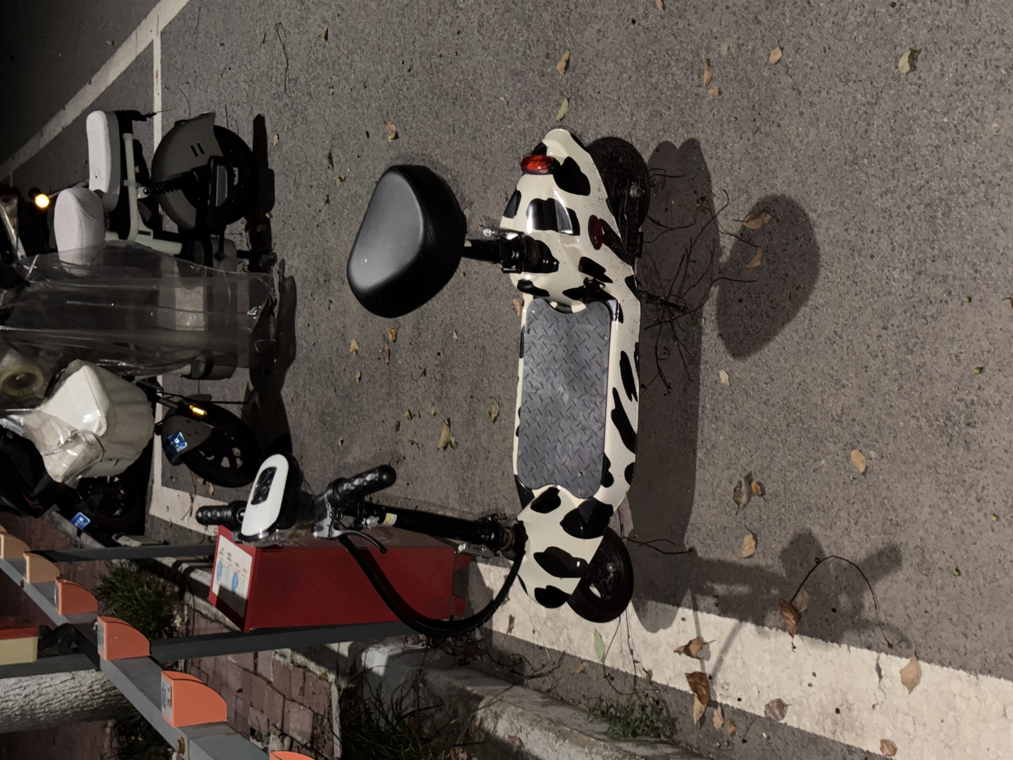 A cow-print electric scooter is parked on an asphalt surface next to a red charging station and a metal pole with orange charging ports. The scooter has a black seat and a patterned deck. In the background, another electric scooter with a white seat and a transparent shield is visible, along with a tree and some fallen leaves. The scene appears to be a public charging area in Jiangjin, China, likely in the evening or at night due to the lighting conditions.