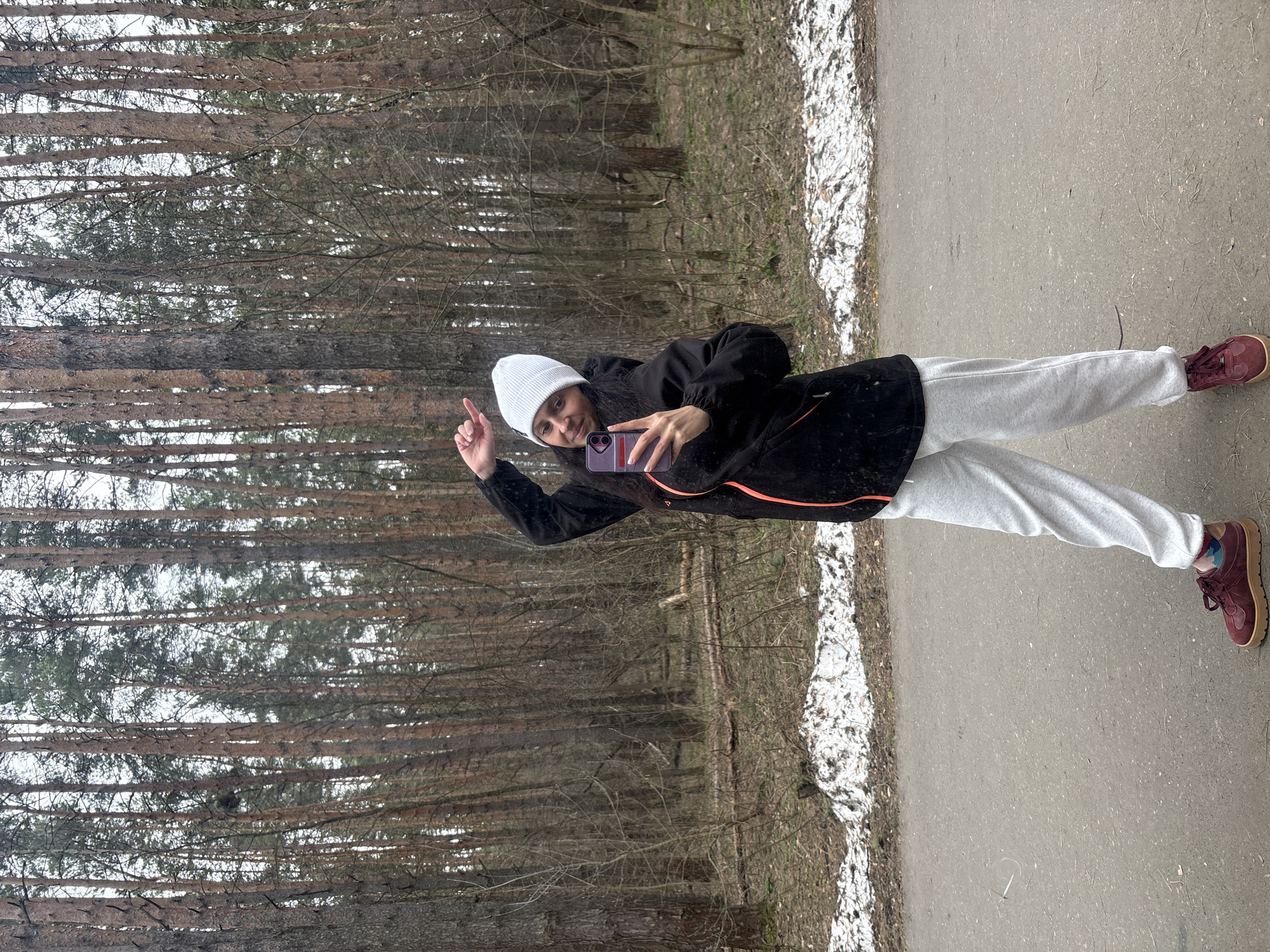 A person is taking a selfie on a path in a wooded area. The person is wearing a white beanie, black jacket with an orange stripe, gray sweatpants, and maroon sneakers. They are holding a purple phone and making a peace sign with their left hand. The background shows tall trees in a forest, with some snow visible on the ground and along the edge of the path. The path is paved and appears to be located within a park or natural area. The location is likely in Neftekamsk, Russia.