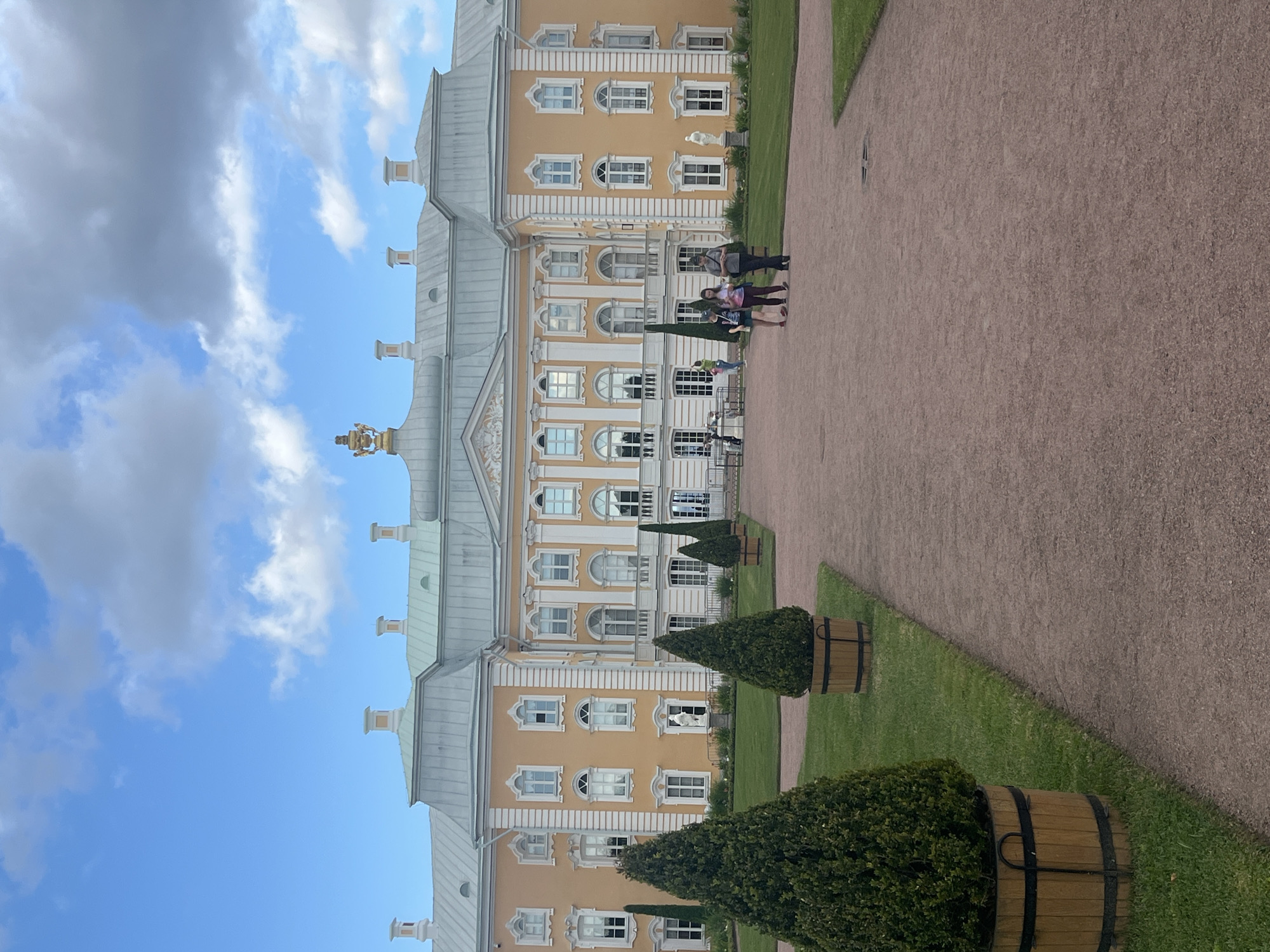 The image shows a grand yellow building with a light green roof and ornate white detailing, typical of the architecture found in Peterhof, Russia. The building appears to be a palace or a large estate. In the foreground, a gravel pathway curves through a manicured lawn, which is bordered by neatly trimmed evergreen trees in wooden planters. Several people are walking on the pathway in front of the building, some of them appearing to be tourists. The sky is a vibrant blue with scattered white clouds. The overall impression is one of a well-maintained historical estate under a pleasant sky.