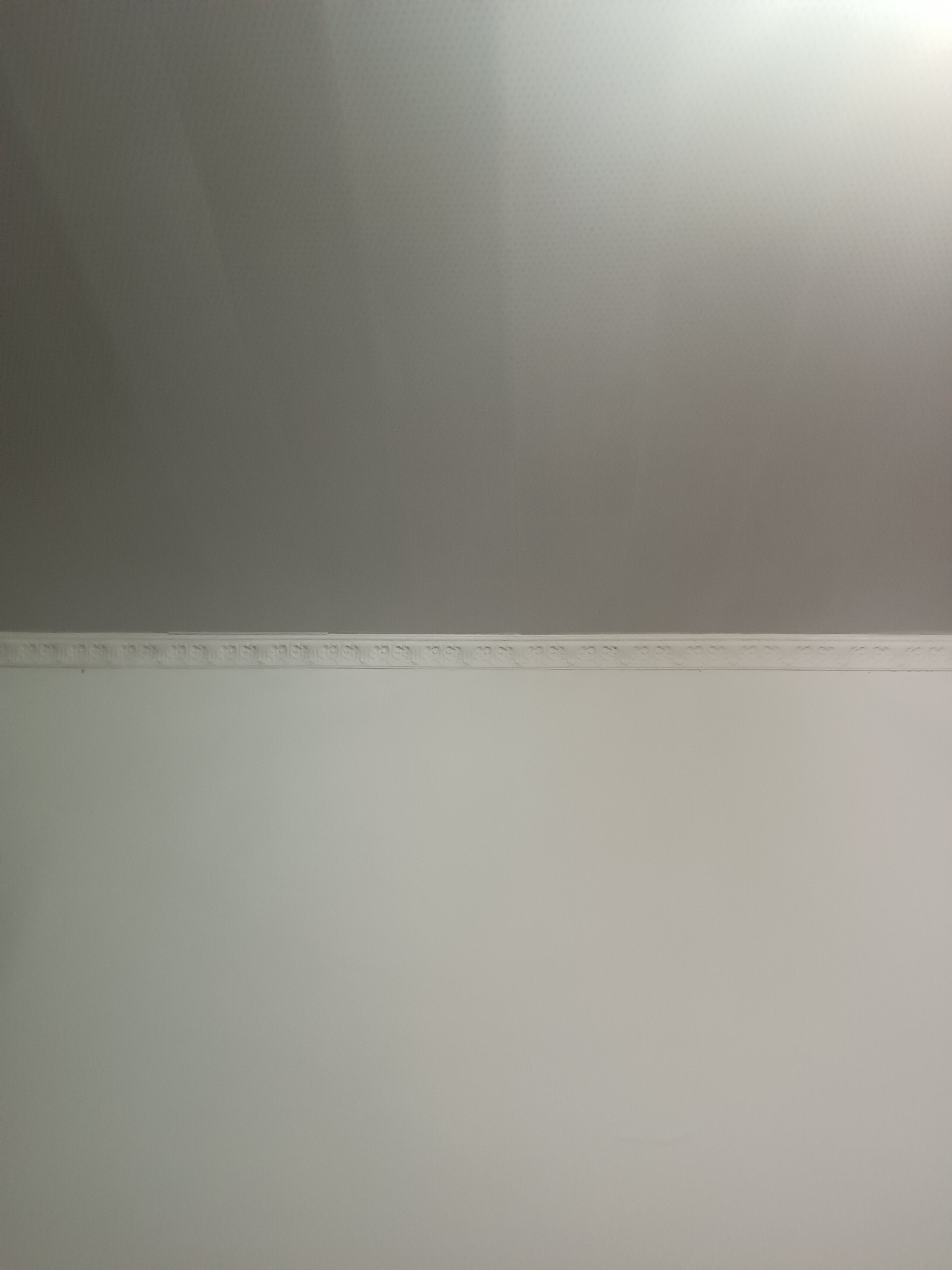 The image depicts a close-up, indoor view of a wall and ceiling, divided by a decorative cornice. The lower portion of the image, occupying approximately two-thirds of the frame, shows a plain, white painted wall. Above this, a decorative white cornice runs horizontally across the image, featuring a repeating, intricate pattern. The upper portion of the image shows a ceiling, which appears to be a light gray color, transitioning to a lighter, brighter area towards the right side, suggesting a light source from that direction. The surface of the ceiling has a subtle, textured pattern, possibly from paint or wallpaper. There are no people or identifiable objects other than the architectural elements of the room. The scene appears to be a generic interior, likely a room in a residential or commercial building, with no specific or discernible details that would associate it with the city of Бидайкөл, Kazakhstan.