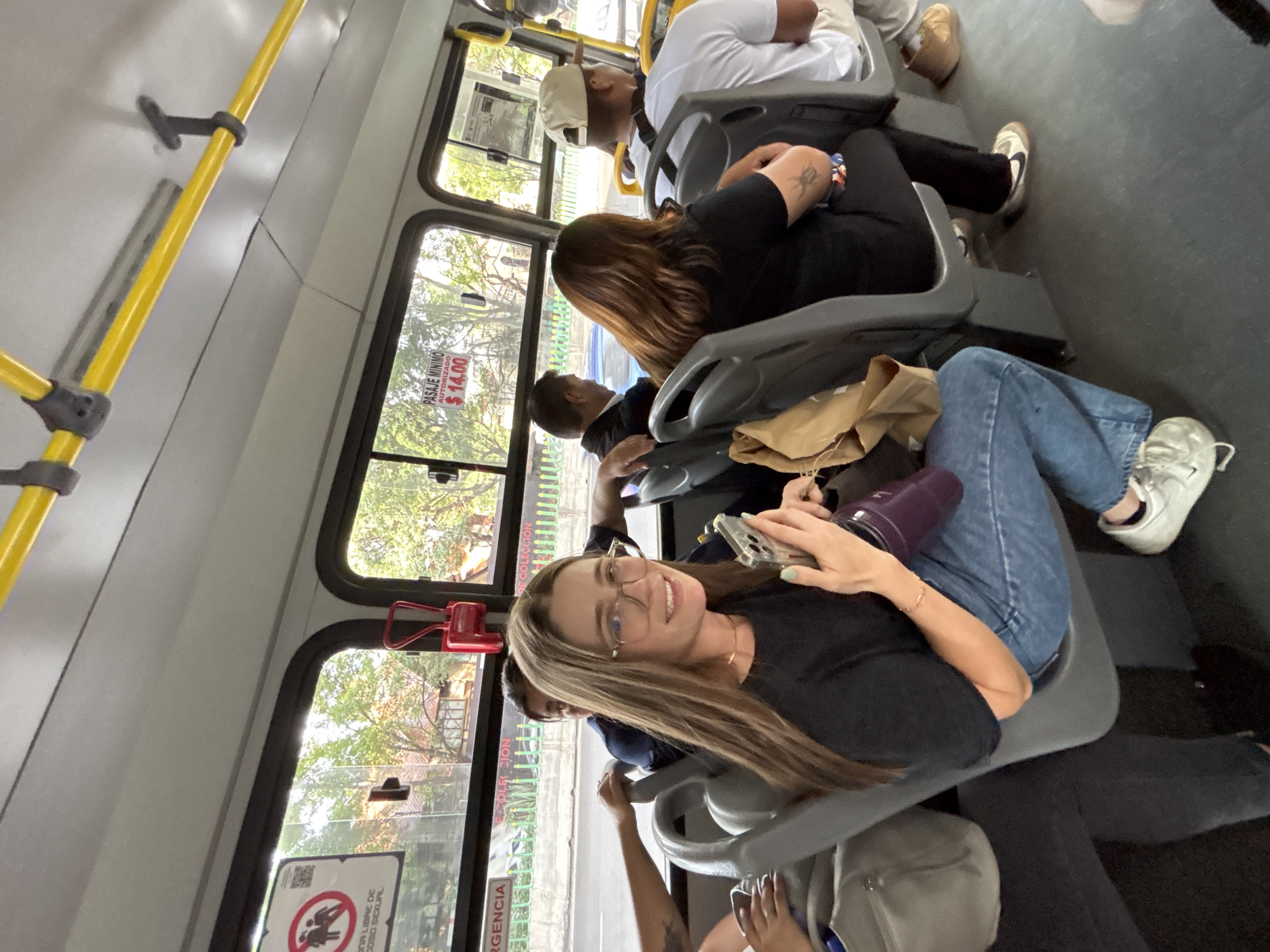The image depicts the interior of a bus, with several passengers seated. The focal point is a woman with blonde hair and glasses, who is looking towards the camera and holding a mobile phone. She is wearing a black top and blue jeans. Other passengers can be seen in the background, including one wearing a white shirt and a cap, and another with brown hair. A yellow handrail is visible near the top of the frame. Outside the bus windows, trees and buildings suggest an urban environment. A sticker with the price "$14.00" is visible on the window, as well as another sticker with a sexual assault warning.