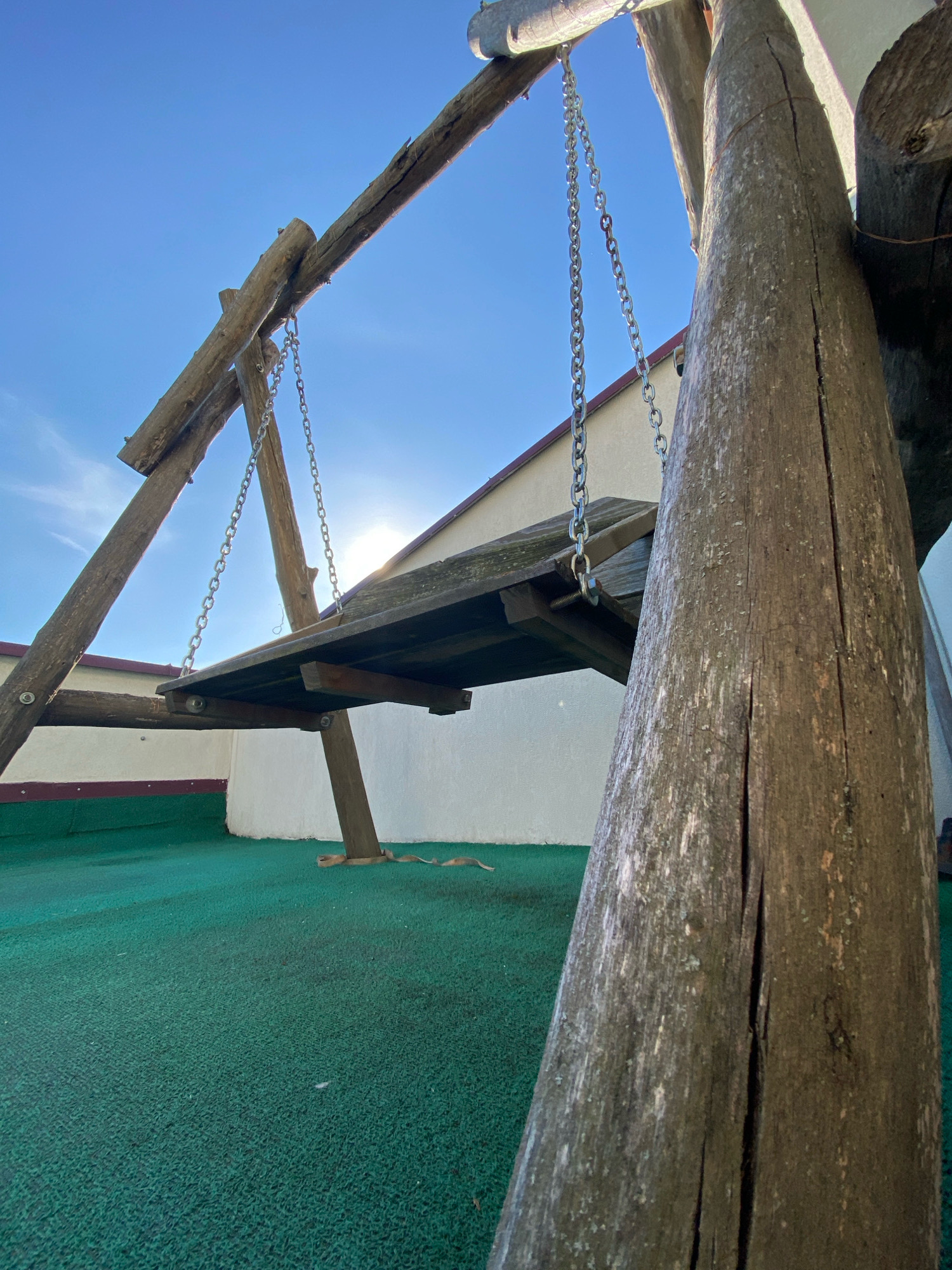 The image depicts a rustic wooden swing set with a wooden plank seat suspended by metal chains. The swing set is constructed from rough, natural logs and branches. It is situated outdoors on a green, textured surface that appears to be artificial turf or rubber matting. In the background, a white building with a reddish-brown roofline is visible under a bright blue sky with the sun shining through. The overall setting suggests an outdoor recreational area or playground. The specific location in Chișinău, Moldova cannot be determined from the visual information provided in the image. There are no people or animals present in the scene, nor any discernible action or interaction. The focus is solely on the swing set and its immediate surroundings.