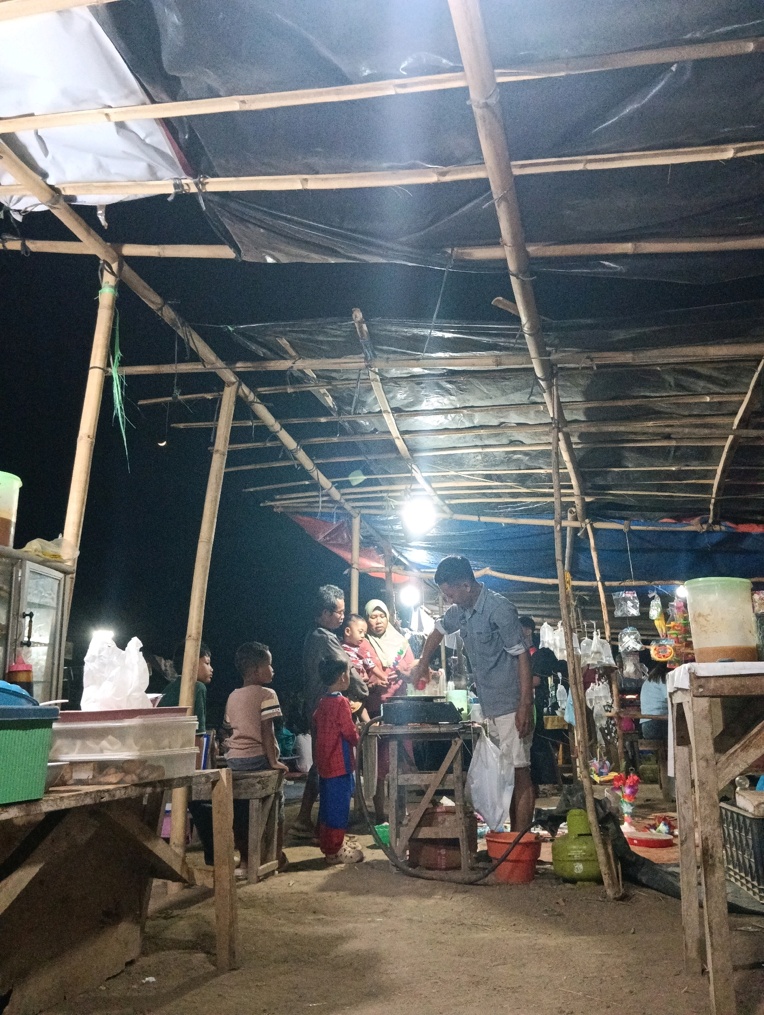 The image depicts an outdoor night market scene in Pematang Jaya, Indonesia. A makeshift stall is constructed with bamboo poles and covered with black plastic sheeting, illuminated by artificial lights. Several people are gathered under the shelter, including adults and children. One man in a light blue shirt is actively engaged in preparing food on a hot plate, while others observe or converse. Items typical of a night market are visible, such as bags of goods, containers, and food preparation equipment. The ground appears to be dirt. In the background, a crescent moon is visible in the dark night sky, suggesting a rural or less developed urban setting.