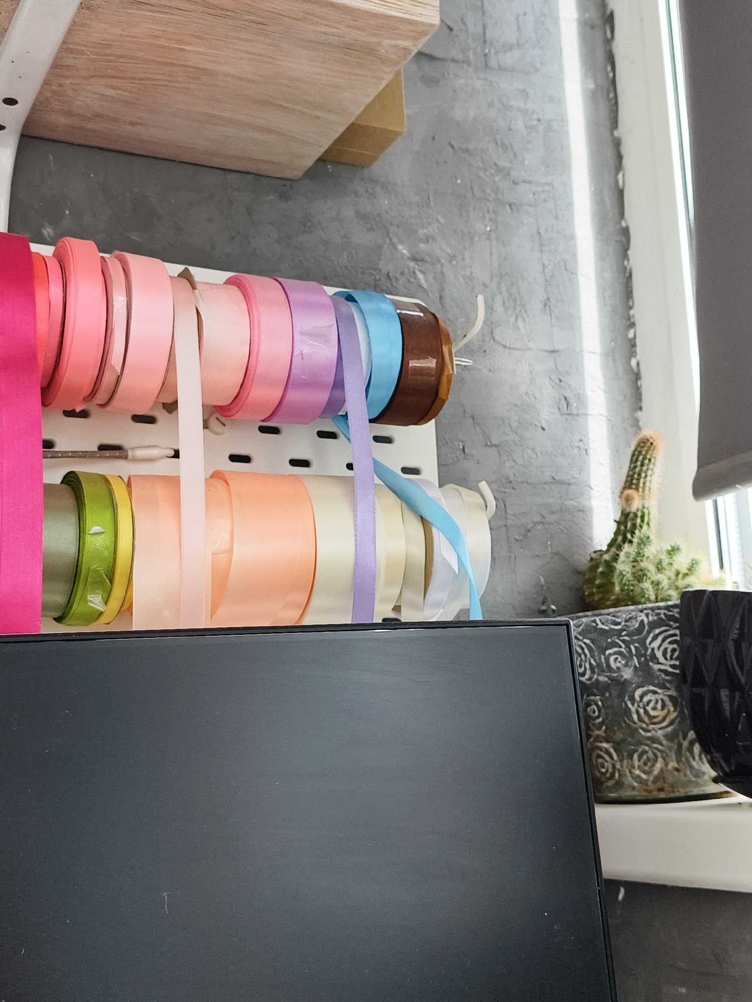 A close-up, eye-level, vertical shot shows a shelf with multiple rolls of colorful ribbon. The ribbons range in color from hot pink, coral, light pink, peach, cream, lavender, and light blue. The ribbons are arranged in two rows on a white pegboard. A cactus in a patterned pot sits to the right of the ribbons. In the foreground, a dark, rectangular object, possibly a monitor or a box, occupies the bottom of the frame. The background consists of a textured gray wall and a window with blinds. The scene is indoors, likely in a craft or flower shop. There is no indication of Volzhsk, Russia.