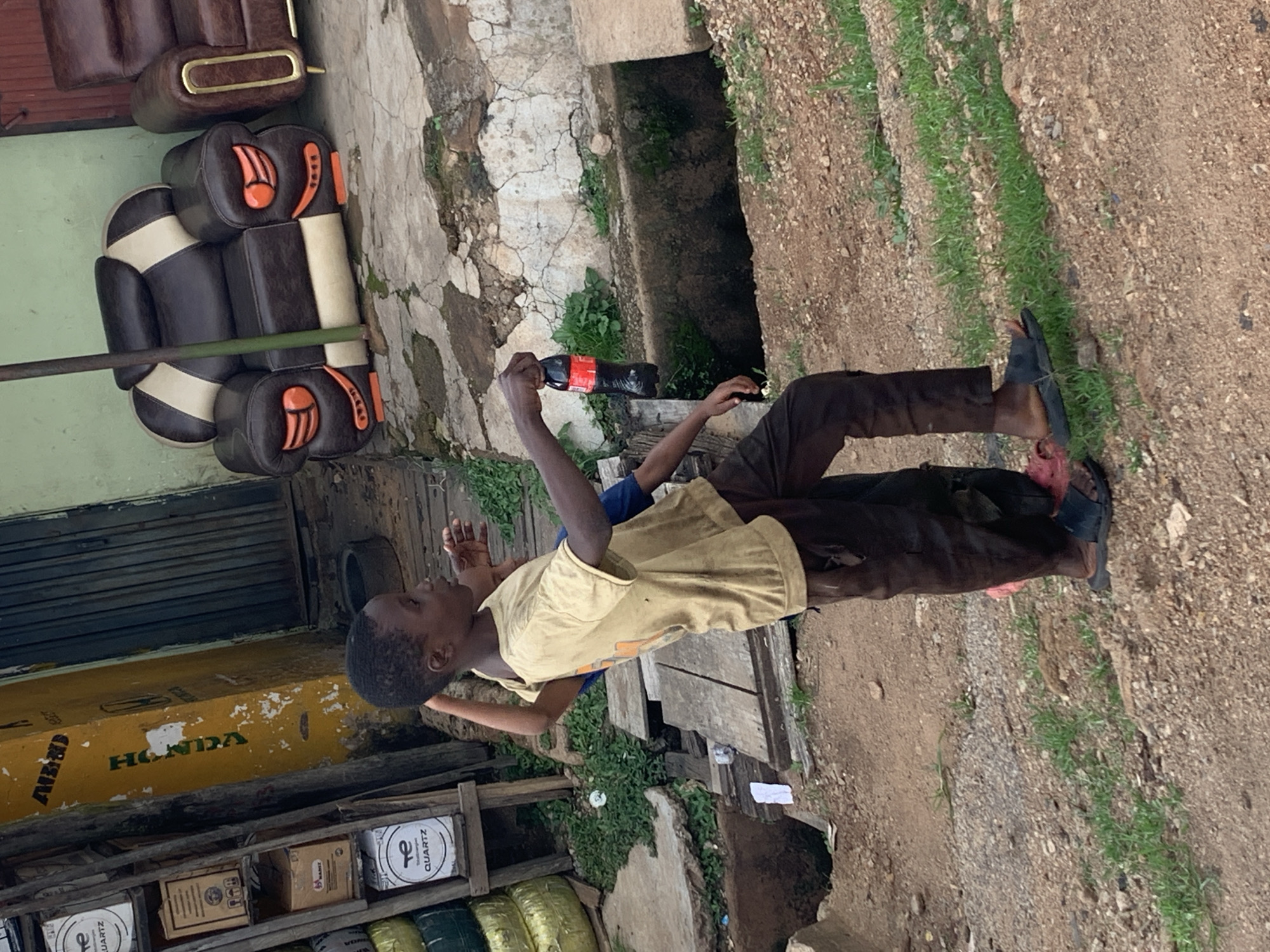 The image shows two young people, likely children, in an outdoor setting that appears to be a developing urban area. The ground is a mix of dirt and gravel with some sparse grass growing. In the background, there is a concrete structure and a drainage ditch. To the left, there are some discarded items, including tires and cardboard boxes. A yellow box with the word "HONDA" is visible on the left side of the image.
One of the subjects, a boy in a light-colored t-shirt and dark pants, is holding up a dark-colored bottle with a red label, which appears to be a soda bottle. He is looking towards the right, and his expression is difficult to discern. The other person, partially obscured, is wearing a blue shirt and is reaching out towards the bottle. The overall scene suggests a casual moment, possibly a pause in play or an exchange. The lighting suggests it is daytime, but the weather is not discernible. The presence of discarded goods and the general state of the surroundings point to a location that may be characterized by limited infrastructure.