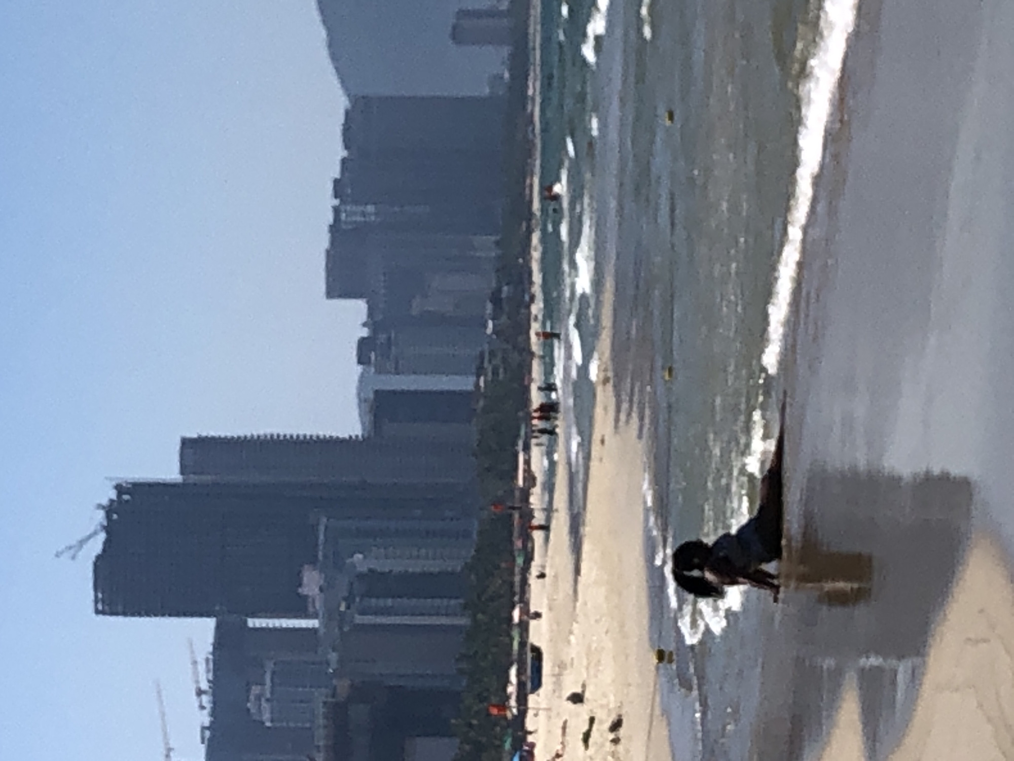 The image depicts a woman sitting on a sandy beach with waves lapping at her legs. She is facing away from the camera, and her dark hair is pulled back into a ponytail. In the background, there is a hazy skyline of tall buildings, indicative of an urban coastal environment. The buildings appear to be modern skyscrapers under construction or recently completed. The ocean is a shade of blue-green, with whitecaps on the waves. The sky is clear and light blue. The overall scene suggests a sunny day at a beach near a developed city. Considering the visual cues of modern high-rise construction along a beach, this location is consistent with the rapidly developing coastal areas of Đà Nẵng, Vietnam, such as the Mỹ Khê Beach area.