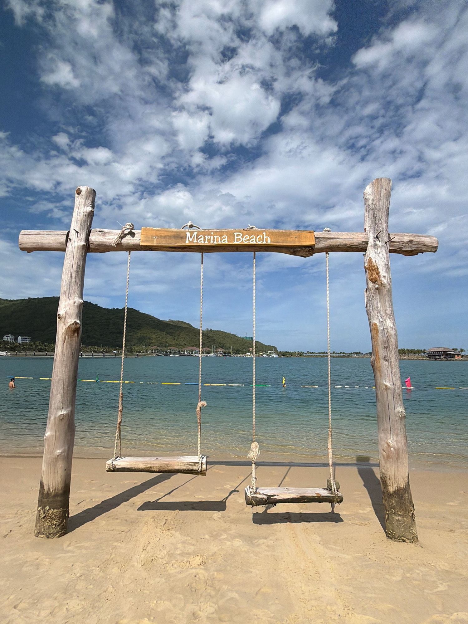 An outdoor, eye-level shot shows a wooden swing set on a sandy beach in Nha Trang, Vietnam, under a bright blue sky with scattered white clouds. The swing set is constructed from rough, weathered logs, with a horizontal beam supported by two vertical posts. Two swings, each with a simple wooden seat suspended by ropes, hang from the beam. A wooden sign with the words "Marina Beach" is attached to the horizontal beam.
The background features the calm, turquoise waters of the sea, with a distant shoreline lined with lush green hills and buildings. Several boats are visible on the water, and a person can be seen swimming near a yellow buoy line. The sandy beach is smooth, with distinct shadows cast by the swing set and swings, indicating a sunny day. The overall scene conveys a peaceful and inviting atmosphere.
