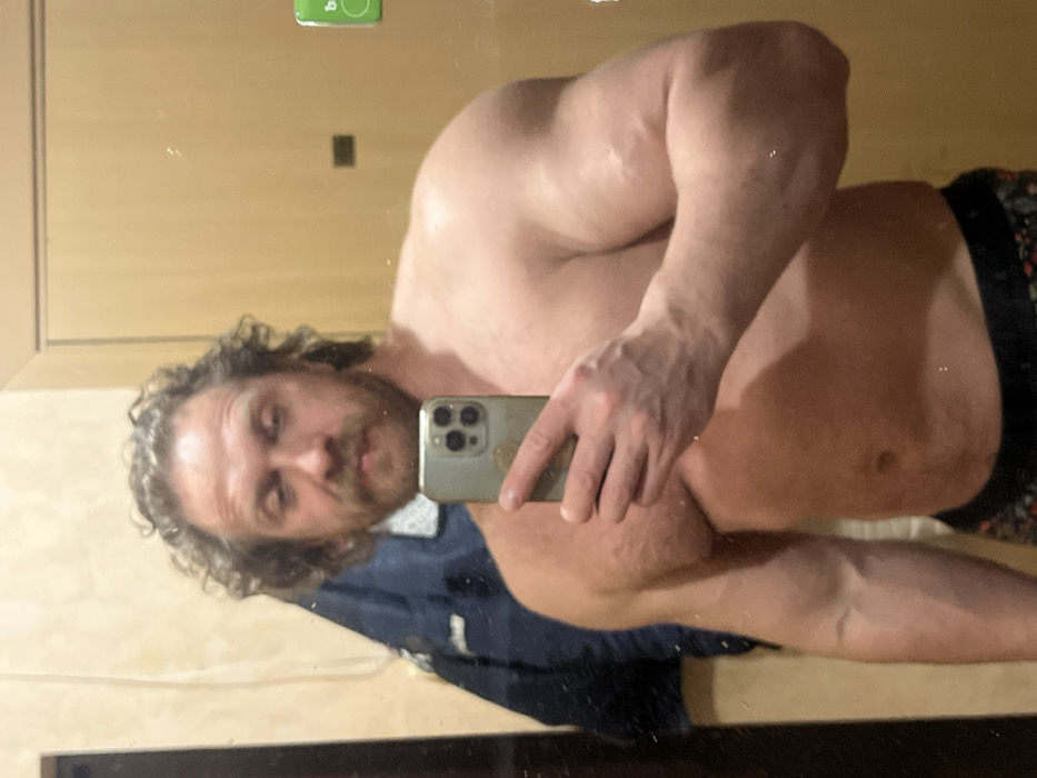 A shirtless man with long, curly hair and a beard takes a selfie in a mirror. He holds a smartphone in his right hand, positioned close to his shoulder. His musculature is visible, especially in his arms and chest. The man is partially visible, as the mirror cuts off part of his body. Dark patterned shorts are partially visible. The background is a simple beige wall with a few small details, possibly switches. A blue garment is visible on the left side of the image.