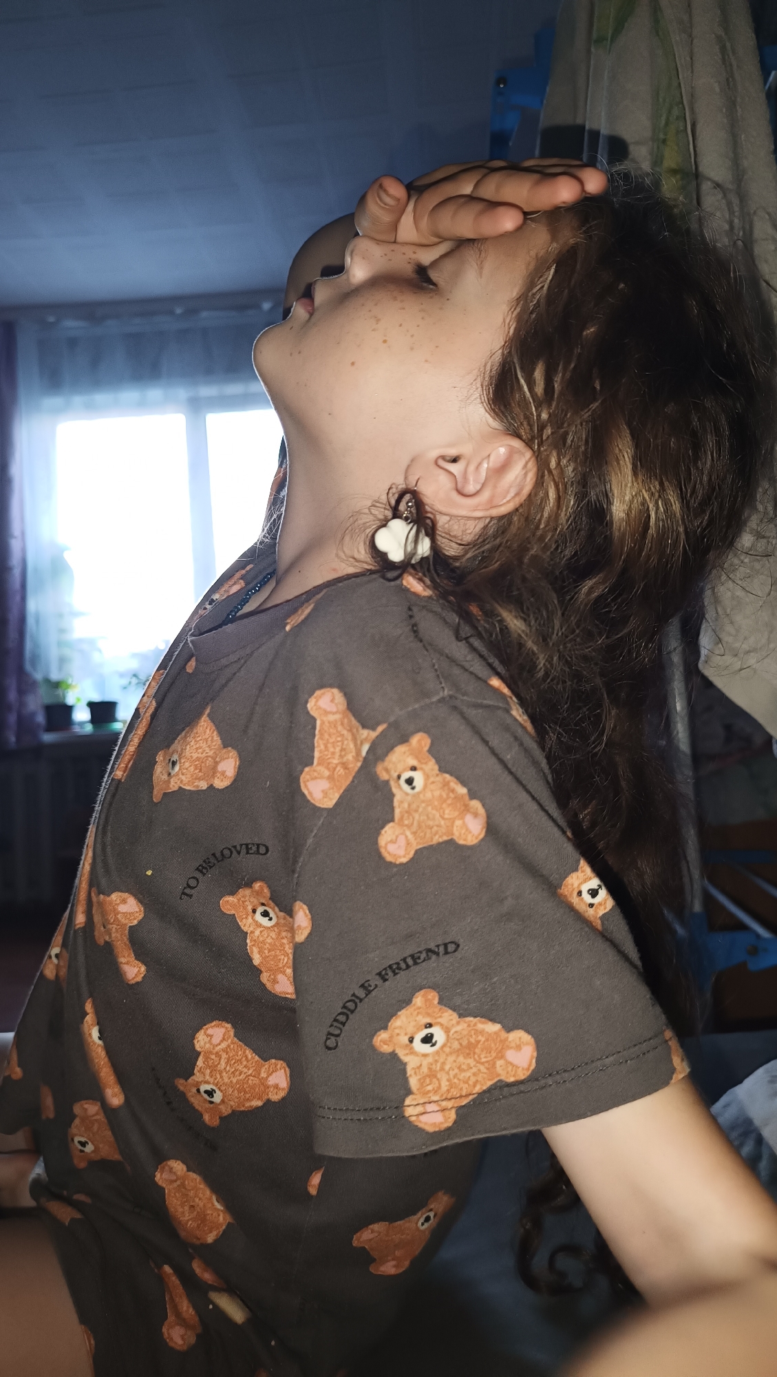 A young girl, likely of European descent, with light skin and freckles, is shown from the chest up, in a profile view facing left. Her right hand is pressed against her forehead. She has long, wavy brown hair that falls over her right shoulder. She is wearing a dark gray t-shirt adorned with a repeating pattern of light brown teddy bears and the words "TO BELOVED" and "CUDDLE FRIEND." She is also wearing a dangling earring in her right ear, which appears to be a white cloud shape with brown strands attached. The background consists of a window with sheer curtains to the left, through which sunlight streams in, and a blurry view of what appears to be an indoor setting with furniture or hanging fabric to the right. The overall lighting suggests it is daytime, with light coming from the left. The image does not contain any visual information to ascertain the specific location within the city of Karsha, Russia.