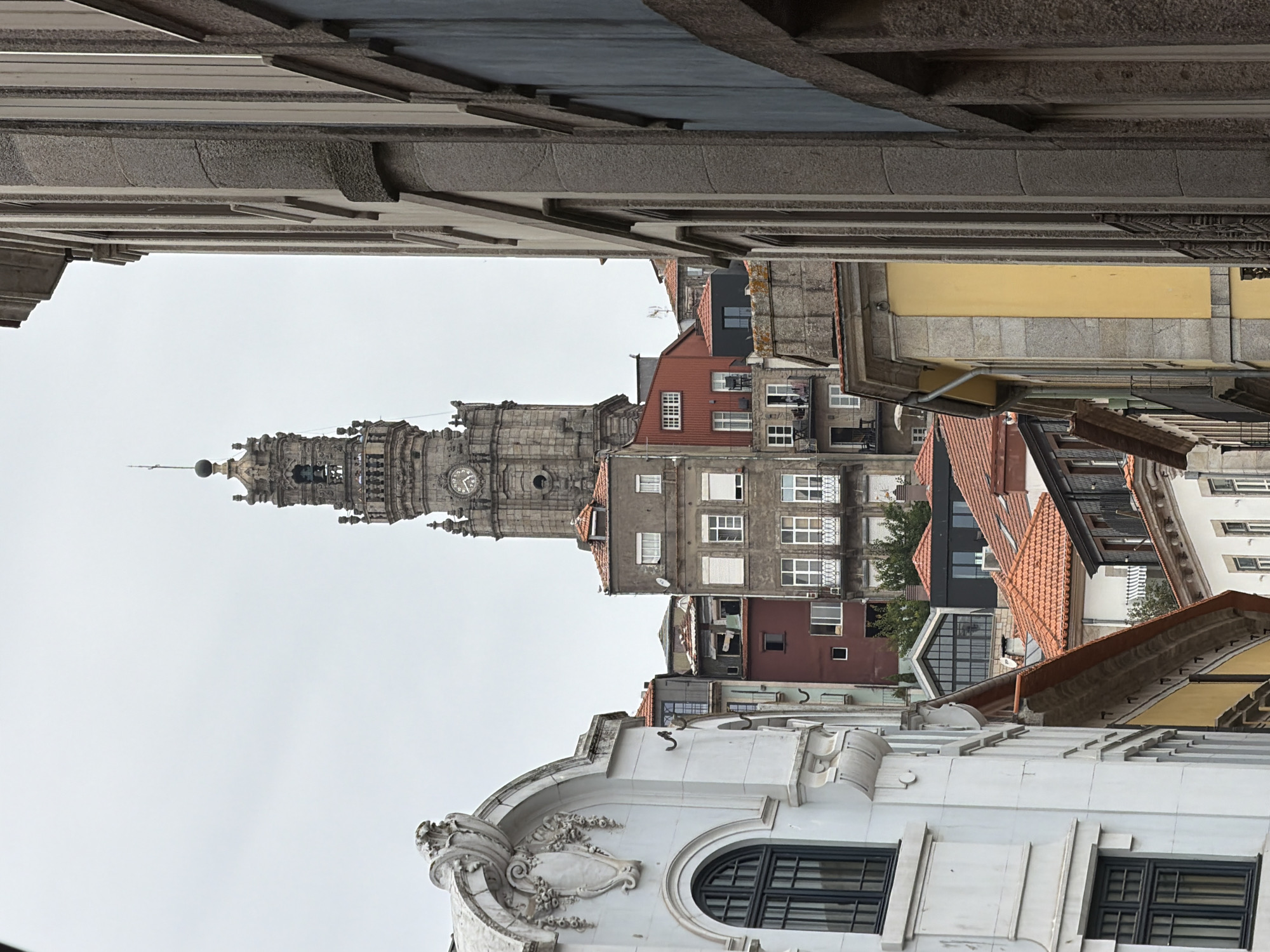 The image depicts a view of the Clérigos Tower in Porto, Portugal, seen from a narrow street. The tower, a prominent landmark, stands tall in the background, characterized by its intricate stone architecture and clock face. In the foreground and midground, various buildings line the street, showcasing a mix of architectural styles and colors.
On the left, a white building with ornate detailing and arched windows is visible. To its right, a cluster of buildings in shades of red, gray, and white are stacked against the hillside, with red-tiled roofs. Some of these buildings have visible windows, balconies, and shutters.
On the right side of the frame, a tall building with a rough stone facade and vertical window openings dominates the view, partially obscuring the scene behind it. This building appears to be much closer to the viewer than the Clérigos Tower.
The sky is overcast, suggesting a cloudy day. There are no visible people or cars in the image, contributing to a sense of quietude or a moment captured without immediate activity. The overall impression is one of urban density, with buildings tightly packed together, typical of historic European city centers. The perspective creates a feeling of looking up and through the urban landscape towards the distant tower.
