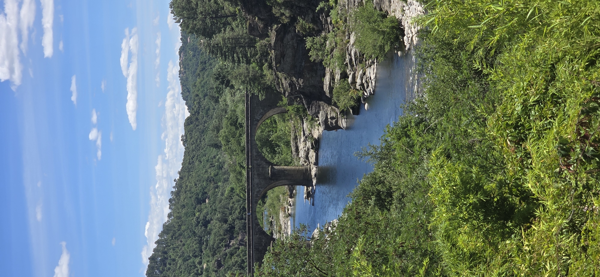 An eye-level, vertical, long shot captures a stone bridge spanning a blue river in a rural, wooded landscape. The bridge, constructed of grey stone, features two large, rounded arches supported by a central pillar. The river flows between rocky banks, with lush green trees and foliage dominating the foreground and background. Hills covered in dense forest rise behind the bridge, and a clear blue sky with scattered white clouds completes the scene. The location is listed as Unknown, Unknown.