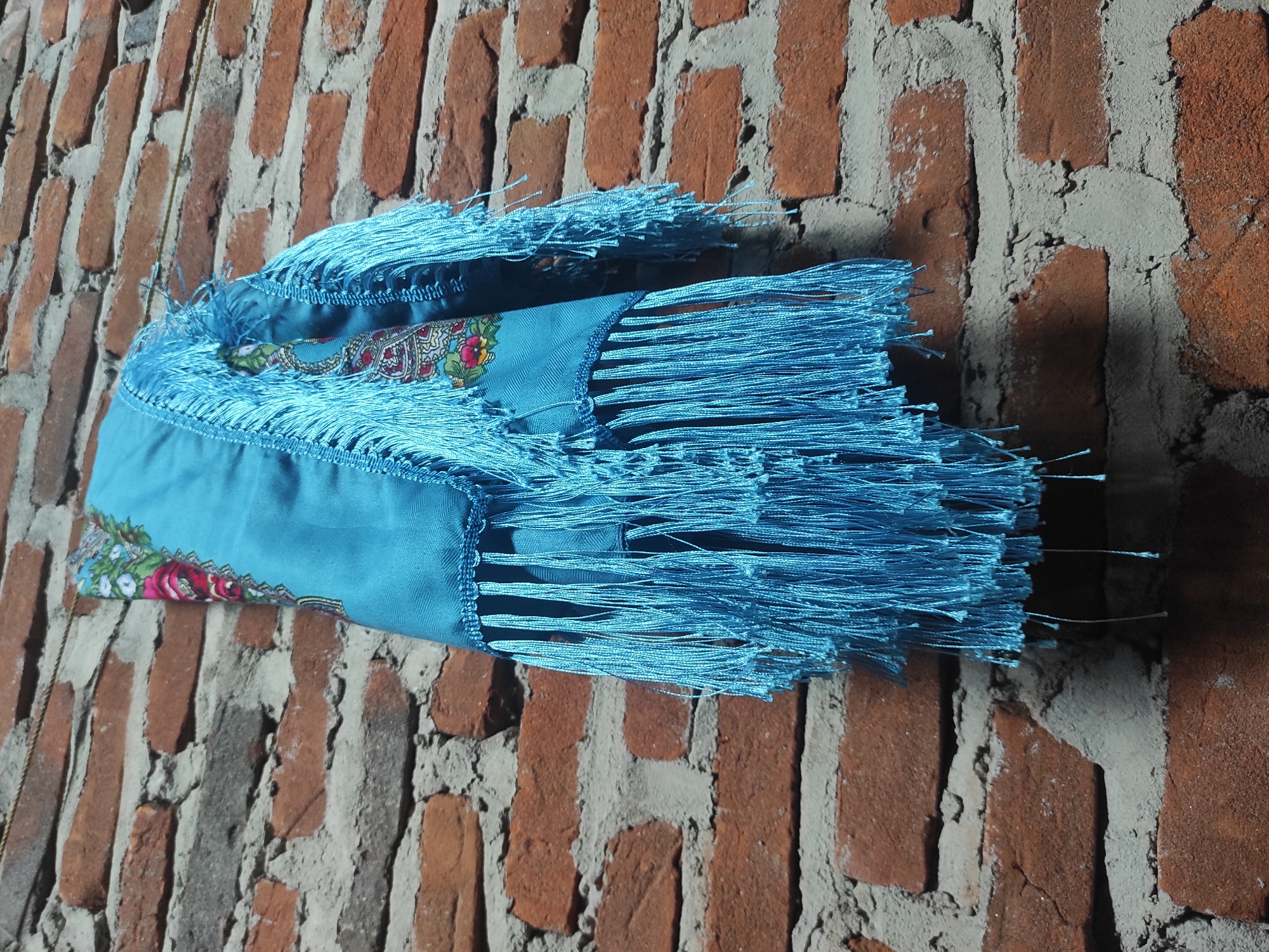 The image displays a folded vibrant turquoise-blue textile, likely a scarf or shawl, leaning against or draped over a rustic brick wall. The fabric is adorned with long, fine fringes of the same striking blue hue along its edges. A visible section of the textile reveals a colorful floral pattern, featuring prominent red and pink roses, green leaves, and other white and yellow floral elements, set against a darker blue or teal background, possibly hinting at a traditional design.
The background is a weathered wall constructed from reddish-brown bricks, which appear aged and have an irregular, rough texture. The mortar filling the gaps between the bricks is light grey, unevenly applied, and shows signs of erosion and wear, contributing to the rustic aesthetic. The bricks are oriented vertically in the frame.
Natural lighting illuminates the scene, casting subtle shadows that emphasize the intricate details of the fabric's fringes and the rugged texture of the brickwork. There is no activity depicted; the image is a close-up, static shot of the textile against its backdrop. No discernible text is visible within the frame. The location context provided for this scene is Lhokseumawe, Indonesia.