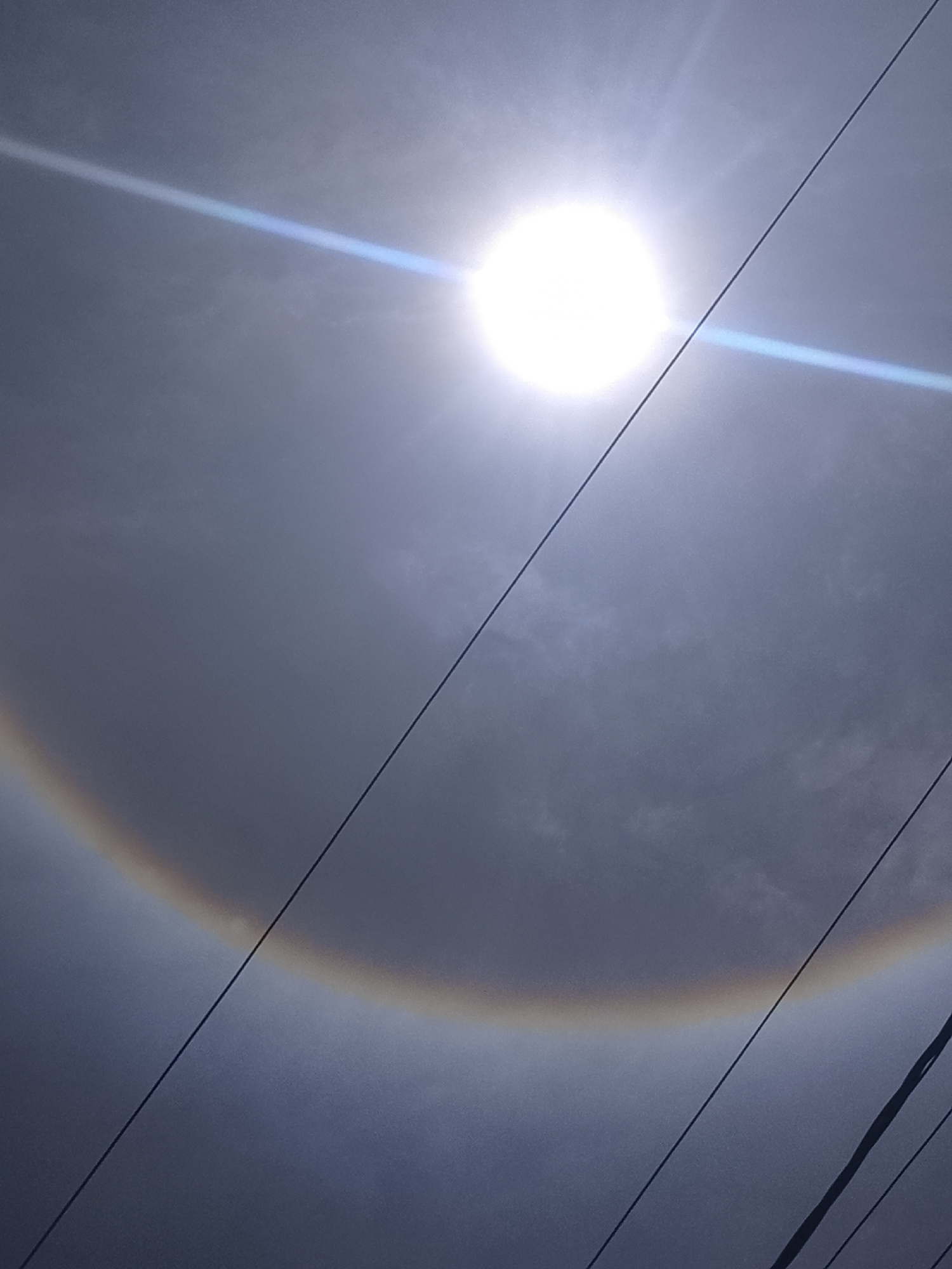 The image shows a 22-degree solar halo in the sky over Altagracia, Venezuela. The sun is visible, surrounded by a bright ring of light. A horizontal light streak is also visible near the sun. Electrical lines run diagonally across the bottom of the frame. The sky is overcast with grey clouds.