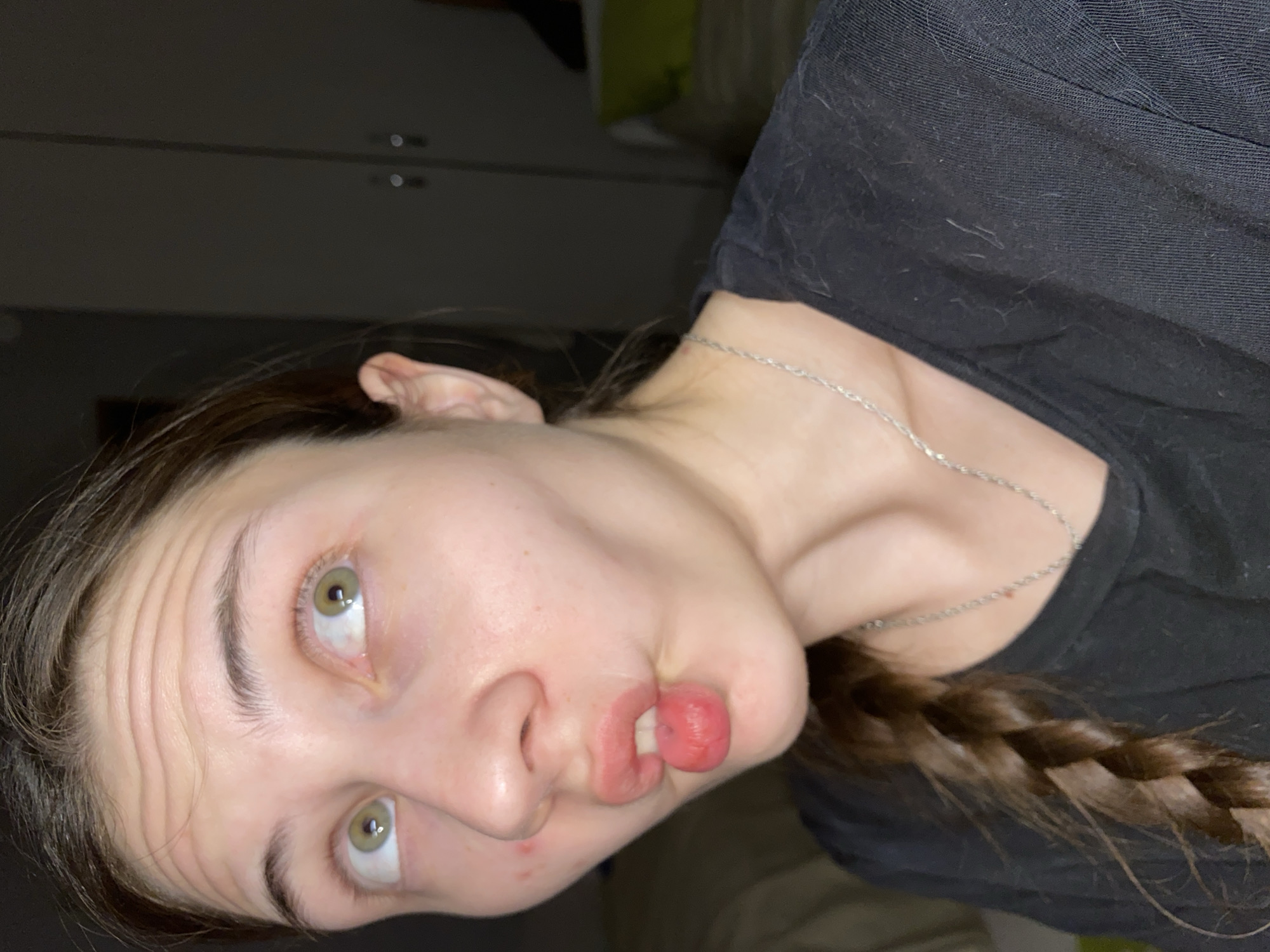 A woman with green eyes and light brown hair, partially braided, wears a black shirt and a silver necklace. She is making a distorted facial expression with her lips protruding. The background shows a light-colored wall or cabinet and a dark, indeterminate space. No specific location in Votkinsk, Russia, is discernible.