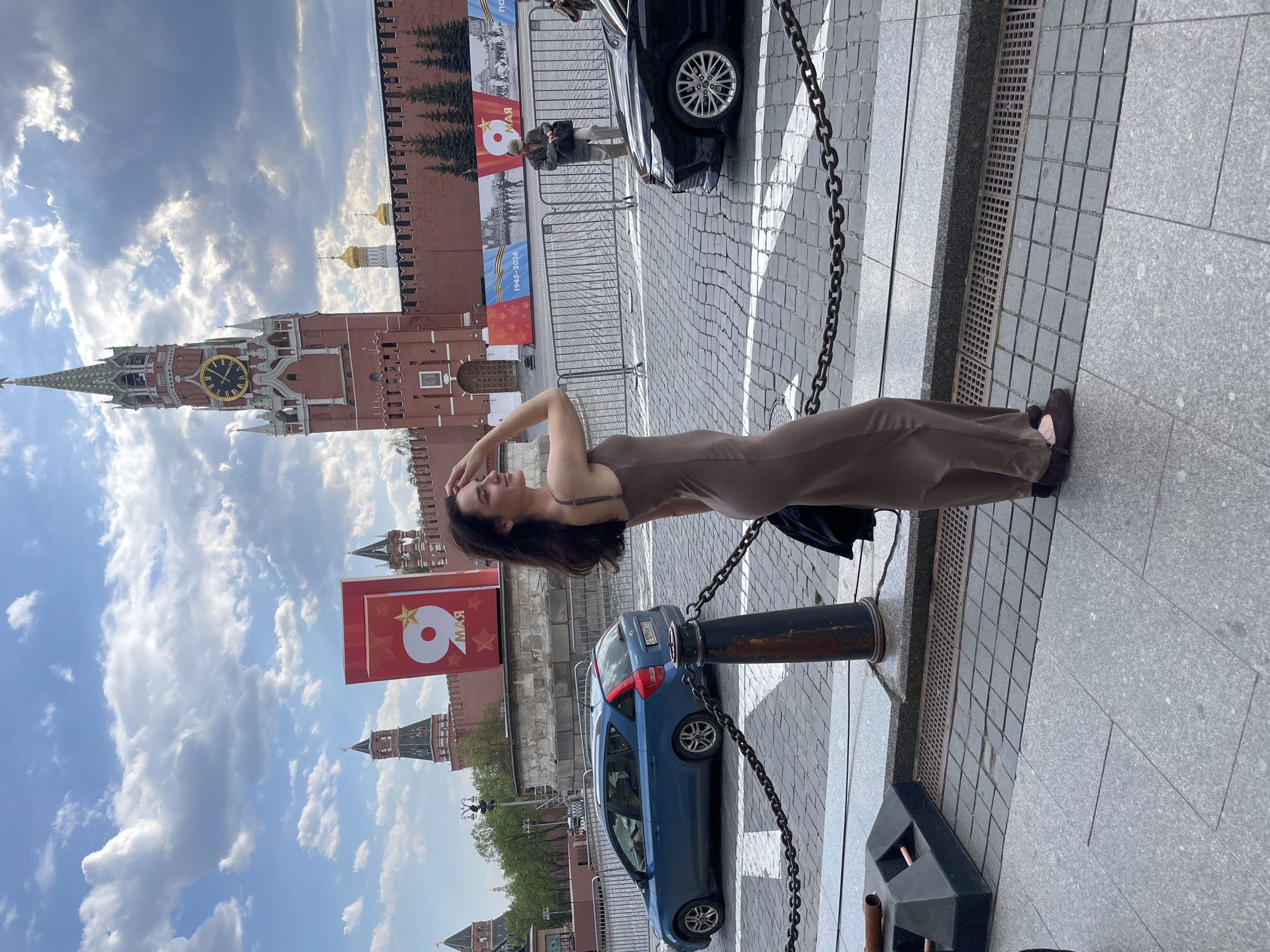 A full-length, eye-level shot shows a woman in a tan dress standing on a gray, bricked area in front of the Moscow Kremlin. She is turned to her right side with her head tilted back, and both hands are on her head. She has dark, long hair. Behind her is the iconic red brick wall and towers of the Kremlin, including the Spasskaya Tower with its clock. A large, red sign with the Russian text "9 мая" (May 9th, Victory Day) is visible. In the background, there is a dark car, a blue car, pedestrian fencing and a gray wall. The sky is mostly cloudy.
