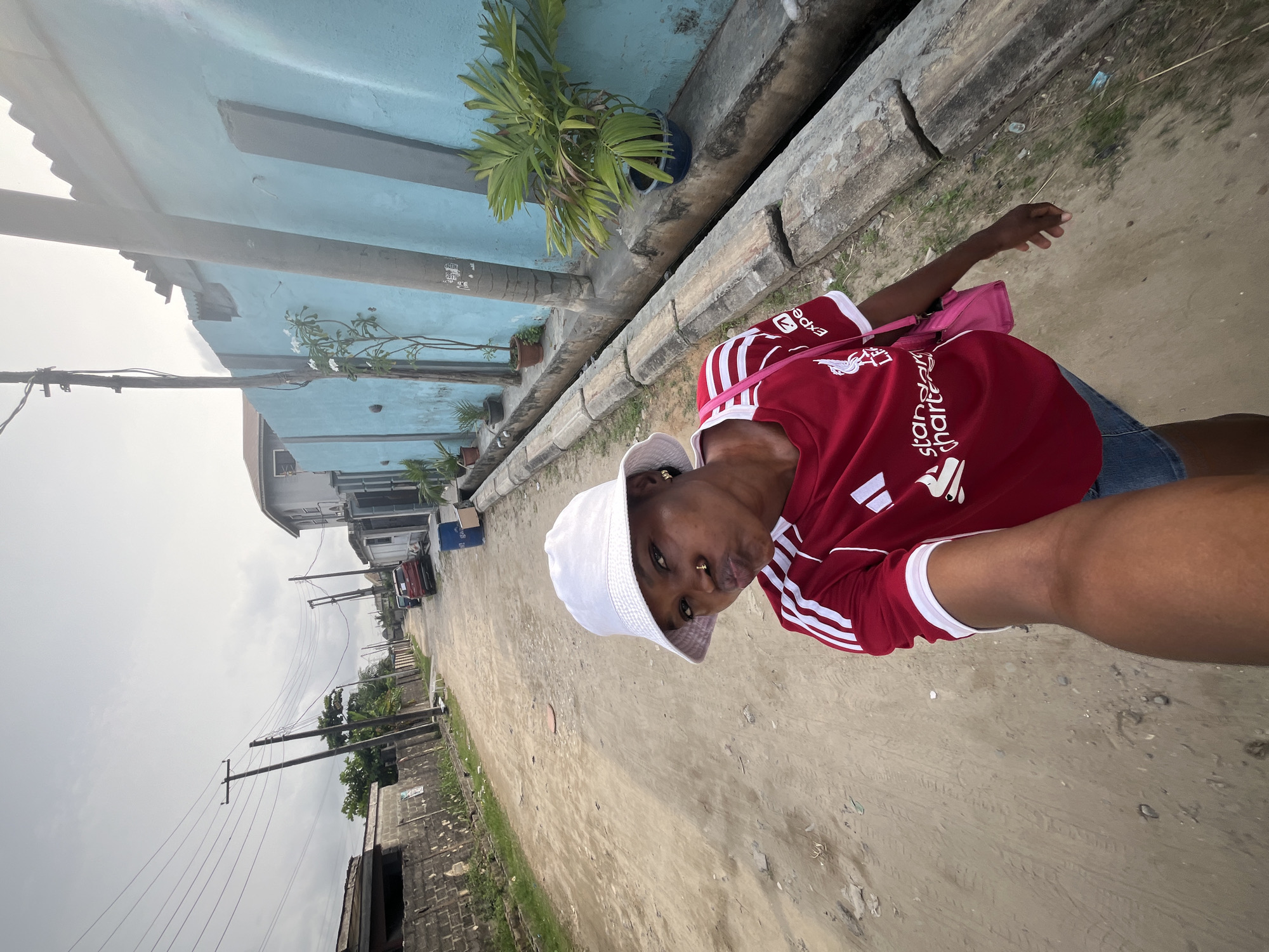 The image displays a person taking a selfie on an unpaved street, with the image rotated 90 degrees clockwise from its natural upright orientation.
**Subjects:** The central subject is a person, facing the camera, wearing a red short-sleeved football jersey with white Adidas stripes on the shoulders. The jersey features the Liverpool FC crest, "Standard Chartered" text, and "Expedia" branding. They are also wearing a white bucket hat, denim shorts, and have a pink shoulder bag. Visible features include a small earring in their right ear and a nose ring. The person is holding the camera with their right hand, part of which is visible in the foreground.
**Scene/Setting:** The setting is an outdoor street or road, which appears to be unpaved, consisting of dirt or sand. On the left side of the street (if the image were rotated correctly), there is a light blue building with a concrete drainage channel or raised curb running along its base. Several potted plants, including palm-like vegetation and flowering plants with white blossoms, are placed along this structure. Further down the street, more distant buildings are visible, along with multiple utility poles carrying numerous electrical wires.
**Activity:** The person is actively taking a selfie, looking directly at the camera with a slight pout on their lips.
**Notable details:** The sky is overcast and hazy, suggesting a cloudy daytime environment. The road surface appears uneven and dusty. The architectural style of the visible buildings and the presence of exposed utility poles are characteristic of certain residential areas.
**Visible text:** Text on the person's shirt includes "Standard Chartered," the Liverpool FC liver bird logo, and "Expedia." White Adidas stripes are present on the sleeves. Some small, unreadable text or posters are faintly visible on the concrete structure next to the blue building.