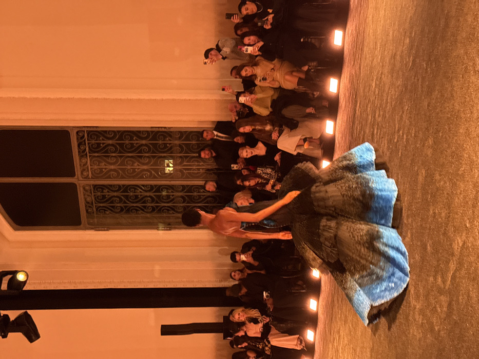 An indoor fashion show is depicted within a grand, historic Parisian building, identifiable by its tall, arched windows featuring elaborate wrought-iron grilles and light-colored walls. The scene is intensely illuminated by a warm, orange artificial light, originating from visible overhead spotlights and ground-level uplights lining the runway.
A model, positioned centrally and seen from behind, walks down a dark, textured runway. She wears a strapless gown with a dark, fitted bodice and a voluminous, tiered skirt that transitions from dark tones to a prominent blue at its lowest tier, trailing behind her on the runway.
To the left and right of the runway, multiple rows of spectators are seated on elevated risers. A significant portion of the audience is actively engaged, holding up smartphones to photograph or record the model. Individuals in the deeper background are observed, some appearing to be event personnel in dark attire. A tall, dark structural element, likely part of the stage rigging, is visible on the far left of the frame.