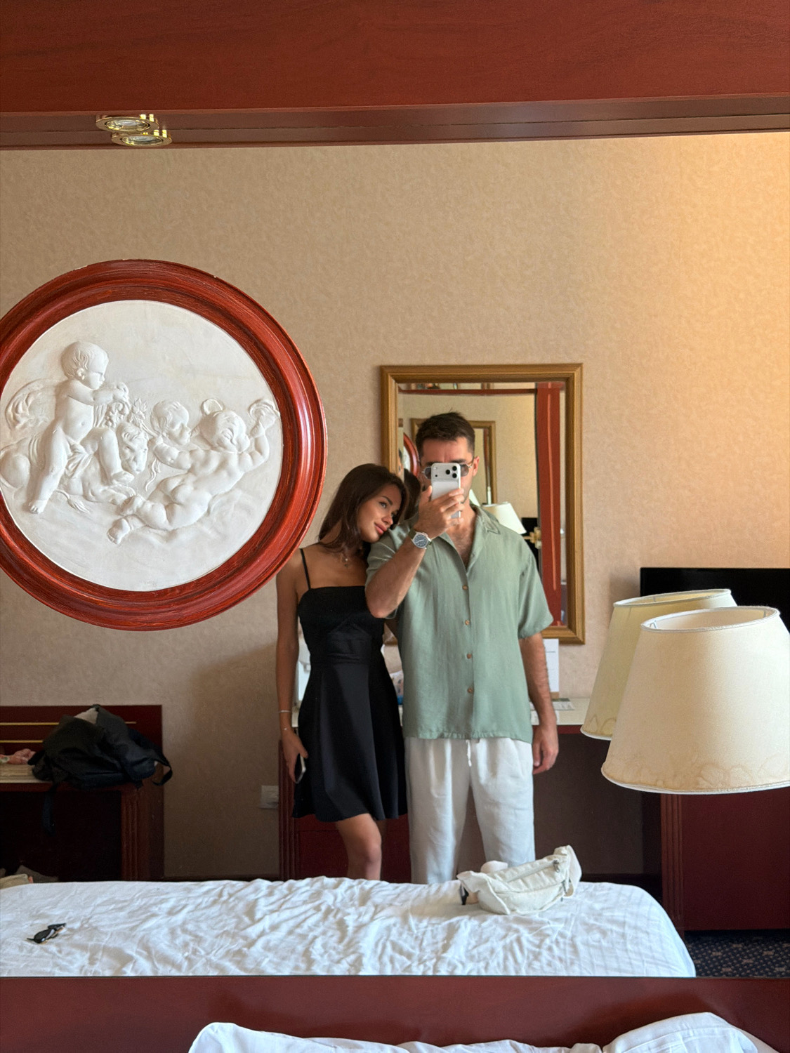 In a hotel room, a young couple poses for a mirror selfie. The woman, on the left, wears a black slip dress. The man, on the right, wears a light green short-sleeved button-down shirt and white pants. He holds up a smartphone to take the photo. In the background, a circular bas-relief sculpture adorns the wall, depicting cherubs. A framed mirror hangs above a dresser. A bed with white linens is in the foreground, and two lampshades are visible to the right. This scene is in Verona, Italy.