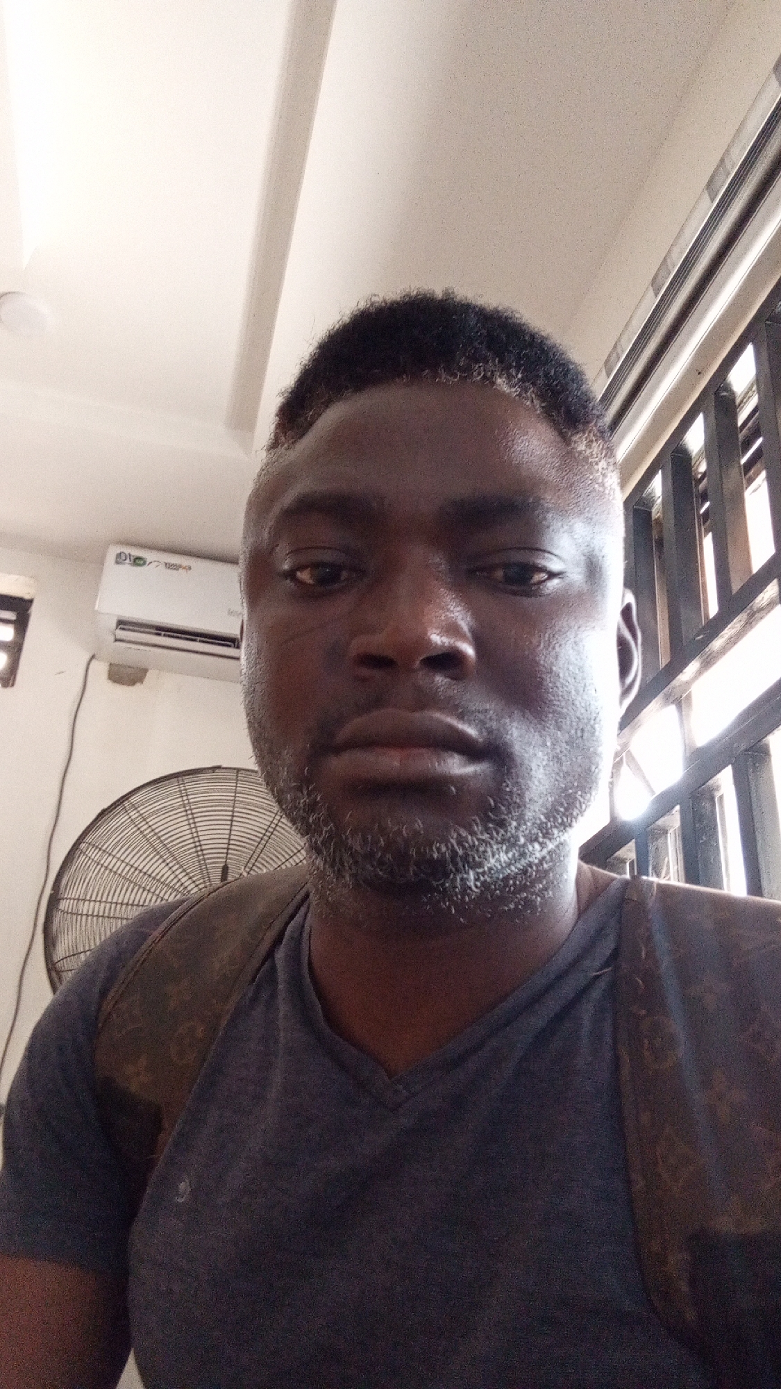 This media file presents a portrait of an adult male, likely a selfie, taken indoors during the daytime. The subject, a man with dark skin, looks directly into the camera with a neutral expression. He has short, dark, curly hair with noticeable grey or lighter strands on the sides and top, and a short beard similarly flecked with grey/white hair. He is wearing a dark grey V-neck t-shirt and a brown backpack with a patterned monogram, resembling a Louis Vuitton design, over his shoulders.
The background indicates an interior setting, possibly in Sardauna, Nigeria, given the presence of cooling appliances. To the left, a white wall-mounted air conditioner unit is visible, bearing the text "Inverter" and a green logo that is not fully legible. Below the air conditioner, a large, black, floor-standing electric fan with a protective mesh guard is positioned. On the right side of the image, natural light enters through a window covered by dark metal bars, suggesting an urban or semi-urban environment. The ceiling is white with some linear architectural features. The man is not engaged in any specific activity other than posing for the photograph.