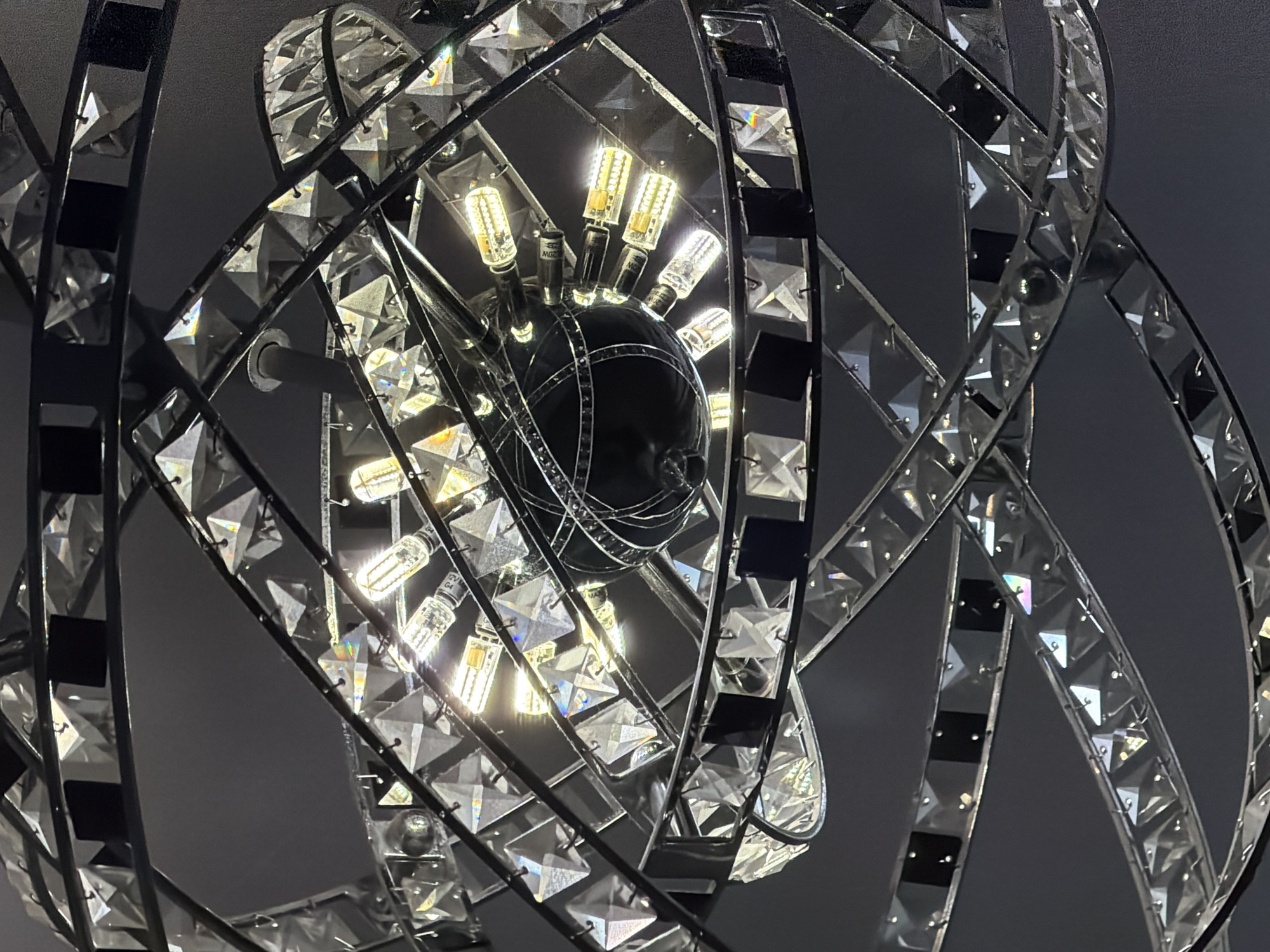 The image shows a close-up of a modern, geometric chandelier with crystal accents. The chandelier features a central spherical element from which multiple small LED light bulbs protrude. The structure is made of dark metal bands that weave and intersect, creating an intricate cage-like design. These metal bands are adorned with numerous clear, faceted crystal pieces that reflect the light from the bulbs, creating a sparkling effect. Some of the crystal facets catch the light at an angle, producing subtle rainbow refractions. Interspersed among the crystals are small black squares, adding a contrasting element to the metallic and crystalline components. The overall impression is one of a luxurious and contemporary lighting fixture. The background is a dark, neutral gray, suggesting the chandelier is likely indoors, possibly in a dimly lit space where its illumination would be a focal point. The lighting in the image is artificial, emanating from the chandelier itself. There are no people or discernible environmental clues to suggest a specific time of day, weather, or explicit location, other than the provided context of Rome, Italy. The image focuses solely on the intricate details and visual appeal of the chandelier. Some of the LED bulbs have faint markings, but no clear, legible text is visible.