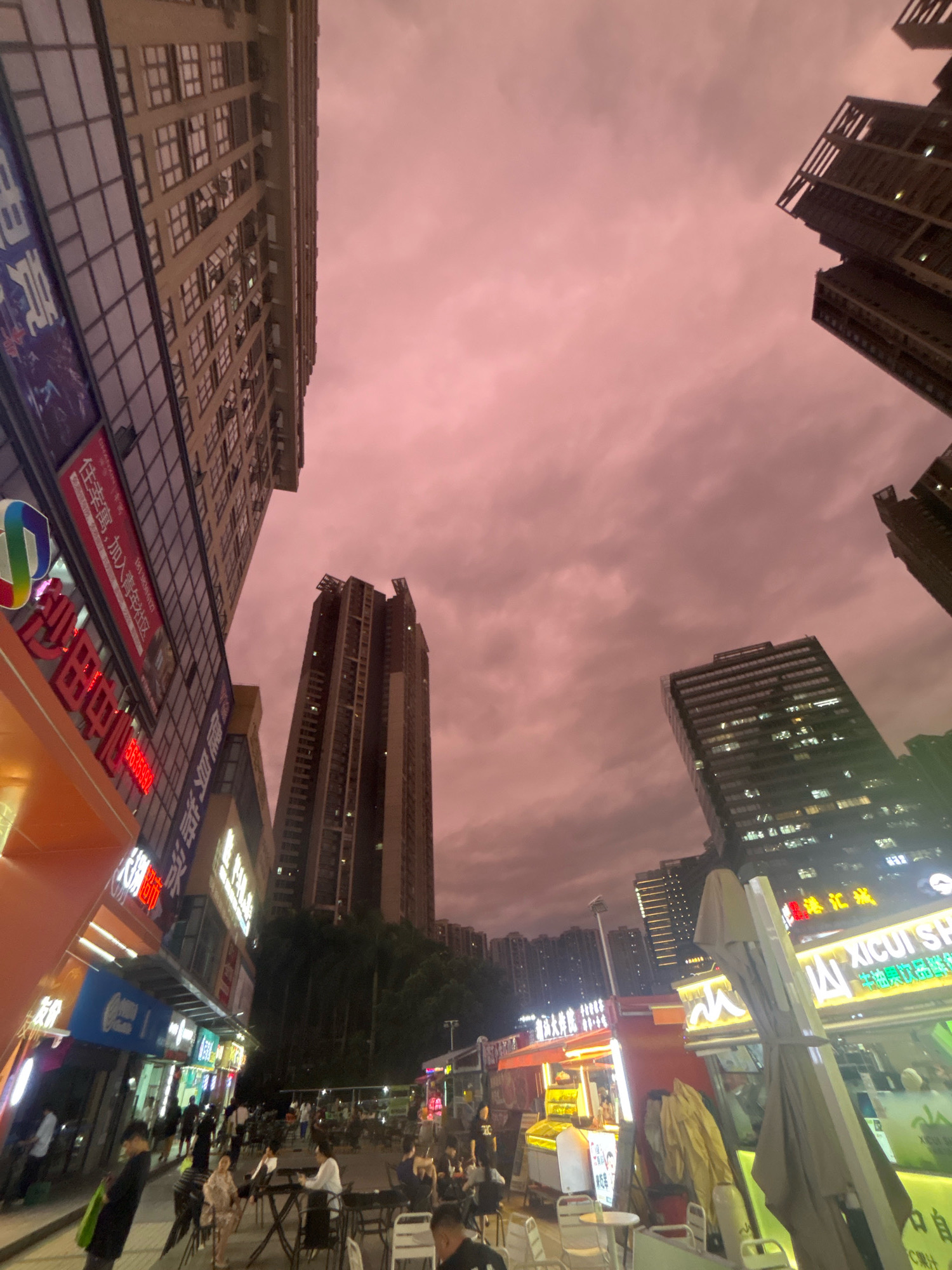 The image captures an outdoor evening scene in Nanhai District, China, characterized by a cluster of modern high-rise buildings and a bustling street-level area filled with food stalls and patrons. The sky is a prominent feature, displaying a dramatic pinkish-purple hue, indicative of sunset or unusual atmospheric conditions, with scattered clouds visible.
Dominating the left side of the frame is a tall, modern building with a facade primarily composed of glass windows and architectural detailing, showing the reflections of the sky. Adjacent to this building, at street level, is a row of brightly lit shops and food stalls, with neon signs displaying Chinese characters. Some of these signs are in red, white, and blue.
In the center and right of the image, other tall residential or office buildings rise, also featuring numerous windows that are illuminated from within, suggesting activity inside. The street level is a vibrant commercial zone. Numerous food kiosks, some with open serving windows and glowing lights, are lined up. Tables and chairs are set up in front of these stalls, with people seated and dining, indicating a lively atmosphere.
A variety of people are visible throughout the scene. Some are walking along the street, while others are seated at tables, engaged in eating or conversation. There are also individuals working at the food stalls. The overall impression is one of a dynamic urban environment with a strong focus on street food and social gathering. The composition, with its low camera angle looking upwards, emphasizes the scale of the buildings against the striking sky.