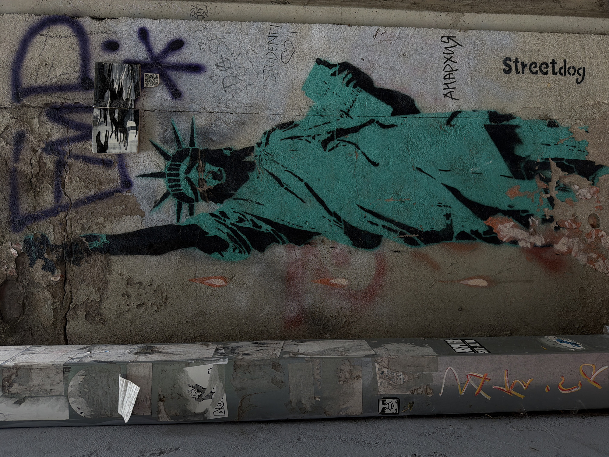 A street art mural of the Statue of Liberty is painted on a cracked, weathered concrete wall in Belgrade, Serbia. The statue is rendered in turquoise and black paint with a stencil-like aesthetic. The wall bears other graffiti marks, including spray-painted symbols and words like "Streetdog" and "ANAPXUA." A rectangular object, likely some type of utility box, runs horizontally along the bottom of the wall, covered in peeling stickers and additional graffiti. The overall environment suggests an urban, neglected underpass or alleyway.
