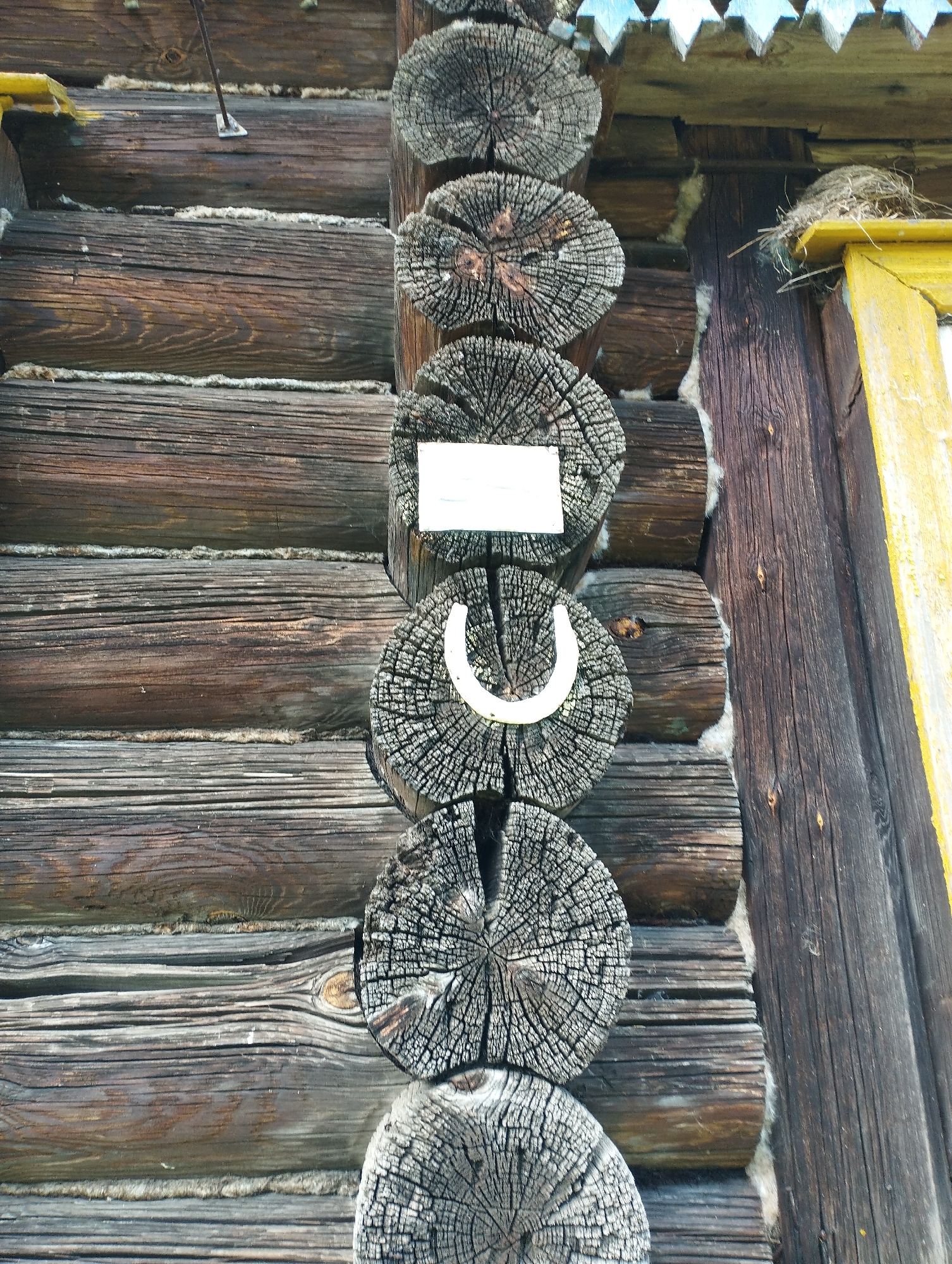 The image shows a close-up, eye-level view of the weathered wooden wall of a rustic building, likely a house, in the village of Shiryaevo (Il'inskiy), Russia. The wall is constructed from horizontal logs that are dark brown and weathered, with visible grain and cracks. Between the logs, a light-colored insulating material, possibly moss or tow, is visible.
Vertically, on the right side of the image, are several cross-sections of logs, also dark and weathered, with concentric growth rings clearly defined. These log ends are likely part of a corner or a structural support.
Prominently displayed on one of the log ends is a white, rectangular sign. Below this sign, another log end features a white horseshoe, positioned upright. Above and to the right of these elements, a portion of a yellow-painted window frame is visible, with a bird's nest constructed from twigs resting on its top edge. The roofline of the building, with its triangular decorative wooden elements, is visible at the very top of the image. The overall impression is one of an aged, traditional rural dwelling.
