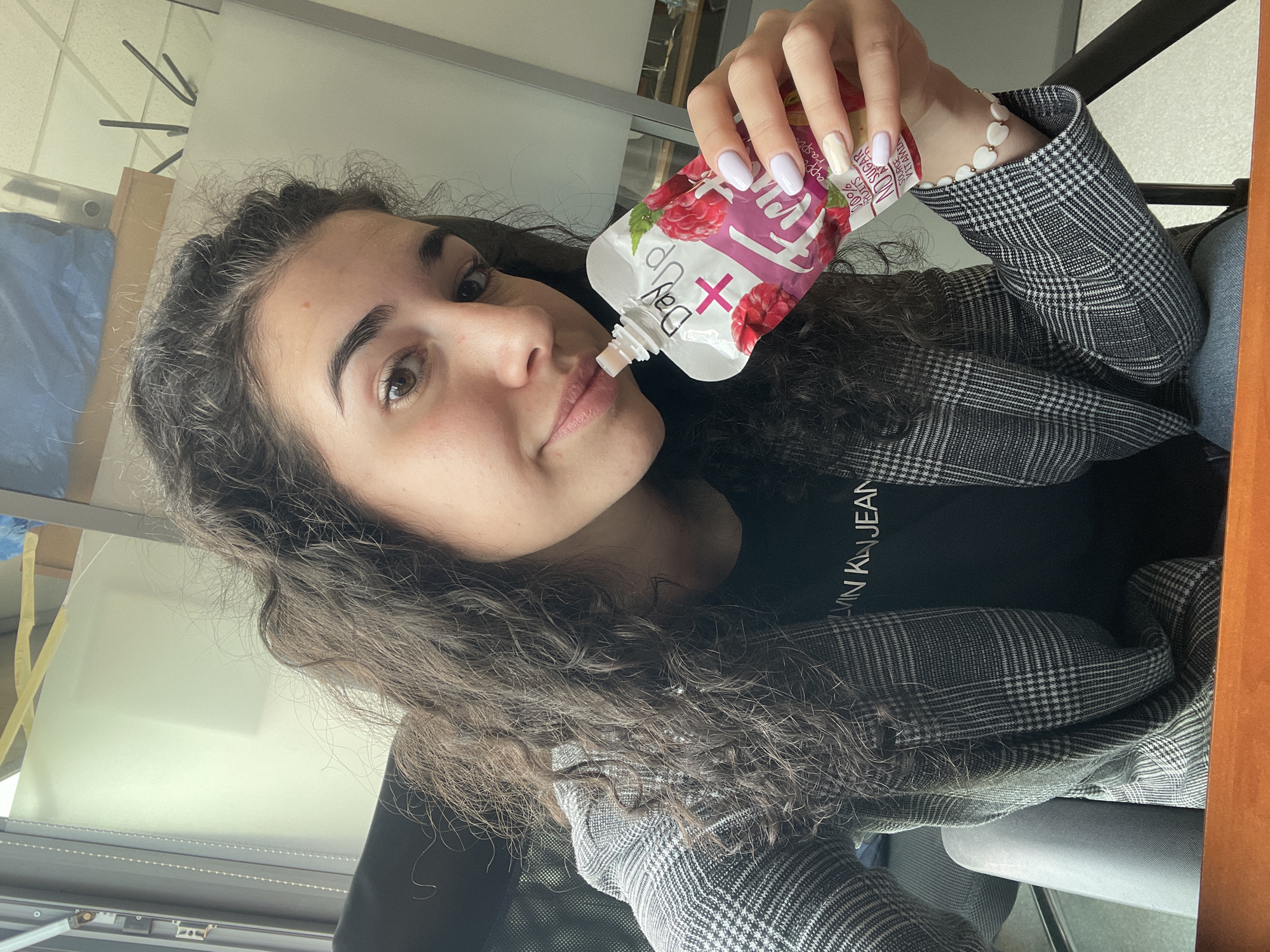 A young woman with long, dark curly hair is drinking a fruit smoothie pouch. The pouch is white with a pink raspberry design and the words "DayUp" on it. She is wearing a black Calvin Klein t-shirt and a grey plaid blazer. She has a white beaded bracelet on her wrist and her nails are painted in light pastel colors. The background appears to be an office setting with a window. There is no information in the image to conclude that the location is in the city of Sofia, Bulgaria.