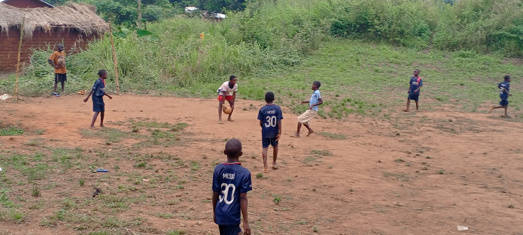 An outdoor scene captures several young boys engaged in a game of soccer on a reddish-brown dirt field. In the background, dense green vegetation, including tall grass and trees, lines the edge of the playing area, and a simple structure with a thatched roof is visible on the far left. The setting appears to be a rural or semi-rural village, consistent with Ngalula, Democratic Republic of the Congo.
Approximately eight boys are scattered across the frame. One boy in the center is holding a light brown, possibly deflated, soccer ball. Two boys, one in the foreground facing away and another further back, are notably wearing identical navy blue jerseys with "MESSI 30" printed in white on the back. A third boy, on the far left, wears a blue jersey with the word "QATAR" partially visible on the front. Most of the children are barefoot, although some sandals or flip-flops are visible discarded on the ground or worn by a few.
On the left side of the frame, two wooden poles serve as improvised goalposts for the game. The boys' positions suggest active play; some are walking or standing, while one on the far right appears to be running. The lighting indicates it is daytime, likely under an overcast sky given the soft, diffused light and lack of harsh shadows. The overall image depicts a casual, informal football match, highlighting the children's simple enjoyment of the game.