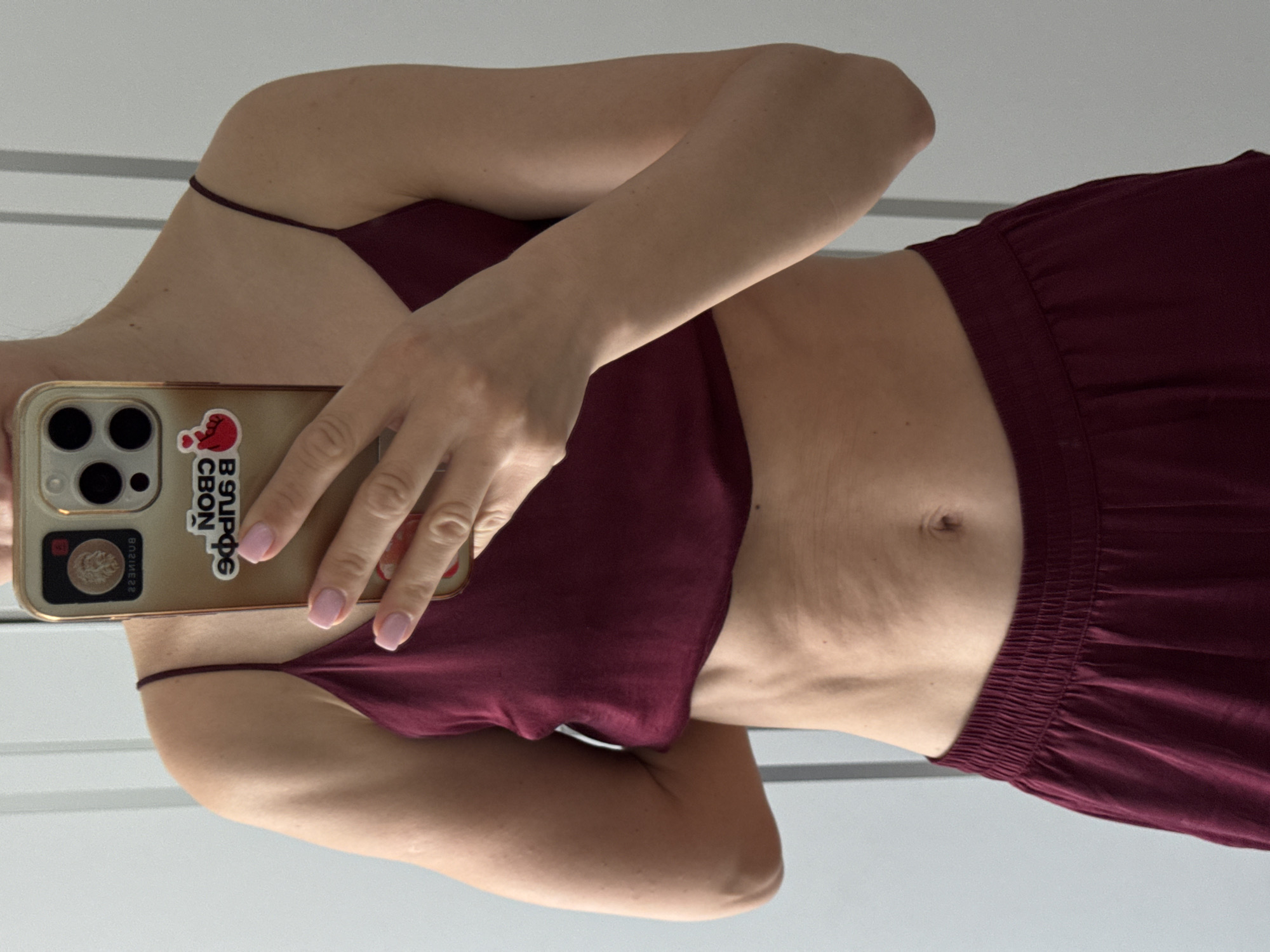 A person, likely female, is depicted taking a mirror selfie indoors in Sochi, Russia. The individual, of fair skin tone, is shown from the chest to the mid-thigh, wearing a matching maroon two-piece set consisting of a spaghetti-strap camisole top that exposes the midriff and elastic-waisted shorts or pants. The left arm is bent, with the hand positioned behind the back, revealing the bicep and shoulder. The right hand holds a gold-colored smartphone, possibly an iPhone, horizontally in front of the body, with the screen reflecting part of the person's head and the room. The nails of the visible fingers are painted light pink. The phone case features two distinct stickers: an upper, round, coin-like sticker depicting a lion's head and the reverse word "BUSINESS," and a lower rectangular sticker with Russian text reading "СВОЙ СБОР" (SVOY SBOR, meaning "MY COLLECTION" or "OWN COLLECTION") and "В ЗНПФЭ" (V ZNPFE), accompanied by a red heart made with fingers. The person's abdomen exhibits natural skin folds, indicating movement or posture. The background consists of a light-colored, possibly white or off-white, wall with vertical architectural lines, suggesting an interior setting with even, diffused lighting.