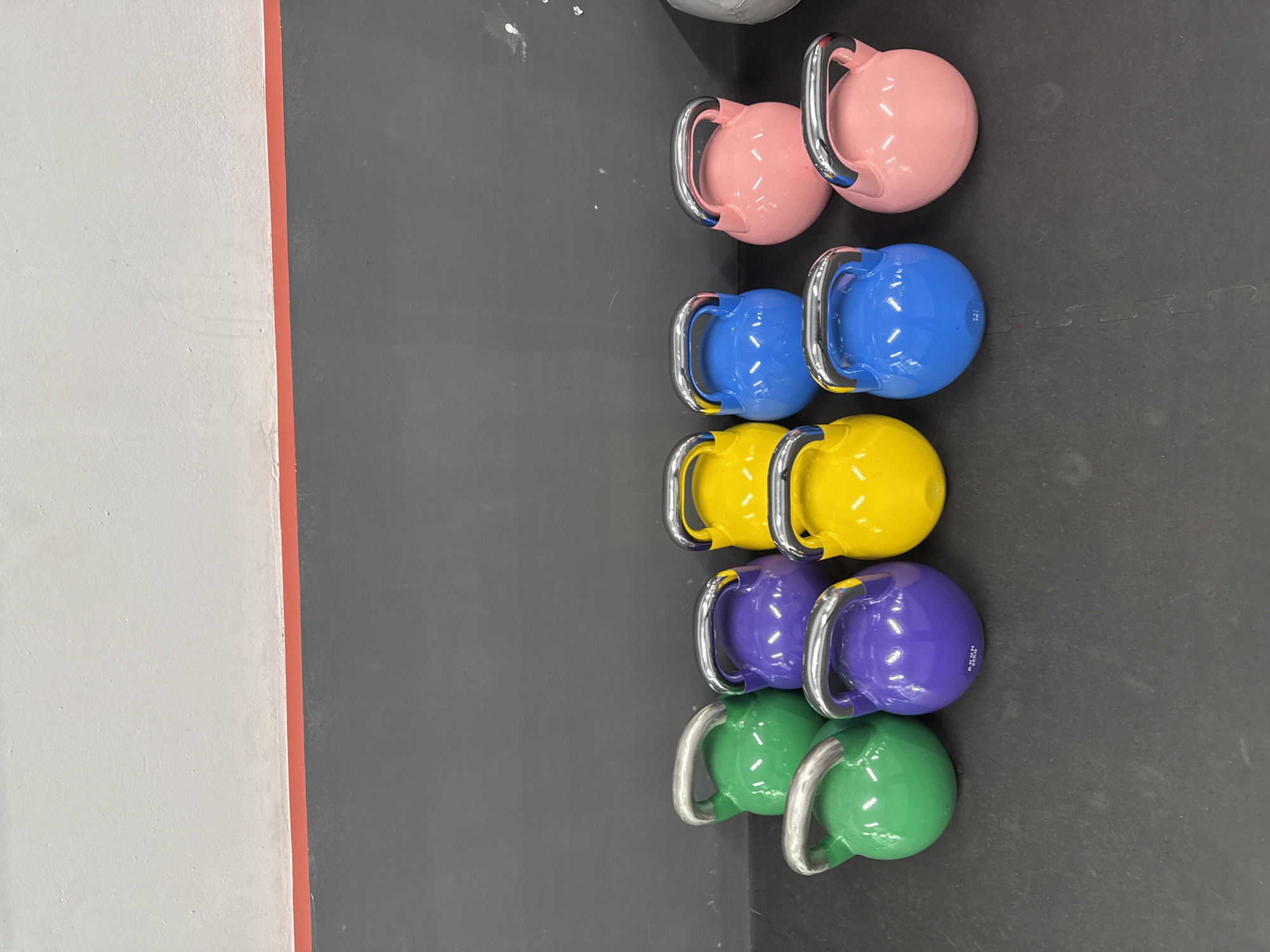 This media file captures a top-down view of five pairs of brightly colored kettlebells neatly arranged on a dark gray, interlocking rubber floor, likely in an indoor gym or fitness facility in Barcelona, Spain.
The kettlebells are lined up in vertical pairs. From top to bottom, the colors are pink, blue, yellow, purple, and green. Each kettlebell features a shiny, chrome-plated handle. The surfaces of the kettlebells are glossy, reflecting the ambient light.
To the left of the kettlebells, there is a light-colored (off-white) wall, separated from the dark gray floor by a thin, reddish-orange vertical strip. The wall has a slightly textured appearance. The dark gray floor shows subtle patterns indicating it is composed of interlocking rubber mats, commonly used in gyms.
A small molded text "MADE IN R.O.C." is visible on one of the purple kettlebells. No people are present, and no specific activity is taking place; the image depicts an organized storage area for fitness equipment.
