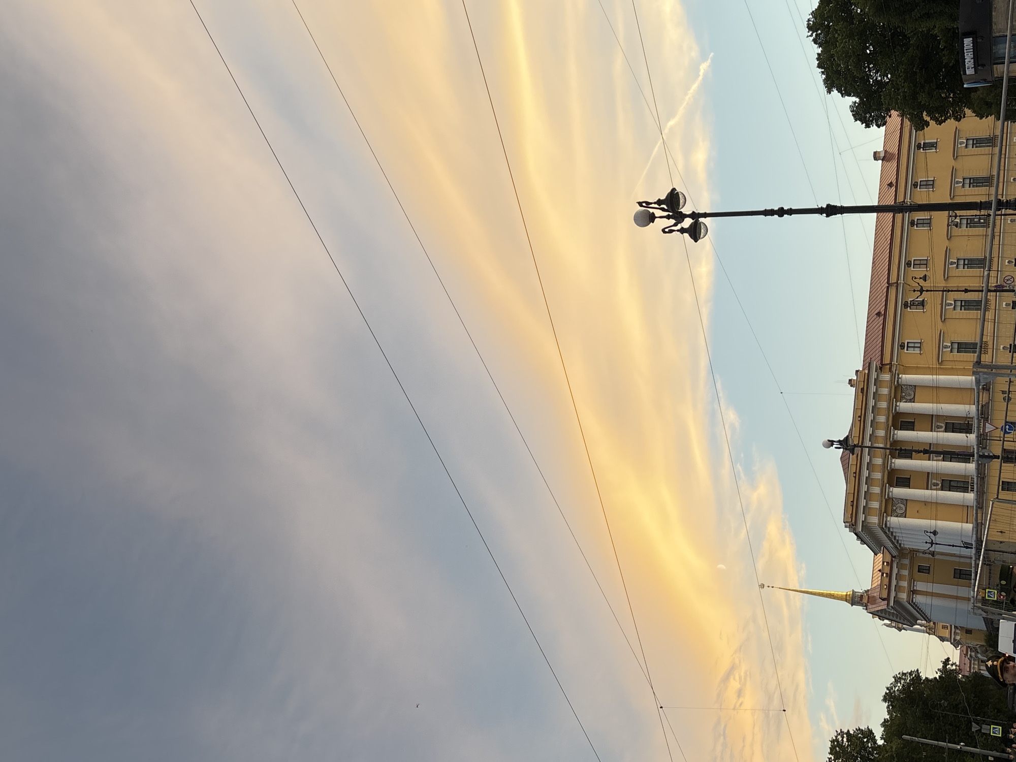 The image captures a view of the Admiralty building in Saint Petersburg, Russia, under a dramatic sky at sunset. The sky is a canvas of soft blues and warm yellows, with wispy clouds illuminated by the setting sun. A thin crescent moon is visible in the upper portion of the sky.
The Admiralty building itself is a prominent structure with a distinctive golden spire, painted in a pale yellow hue with white columns and red roofing. The architecture suggests a historical and grand setting.
In the foreground, a tall, ornate streetlamp with multiple globes stands on the right side of the image, its dark metal contrasting with the bright sky. Several overhead wires crisscross the scene, suggesting the presence of urban infrastructure.
On the left side of the image, the tops of lush green trees are visible, framing the architectural elements. The lower part of the image shows glimpses of people and a street, with traffic barriers and signs indicating a public area. A police officer in uniform is partially visible in the bottom left corner. The overall scene conveys a sense of urban grandeur and the beauty of a city at dusk.