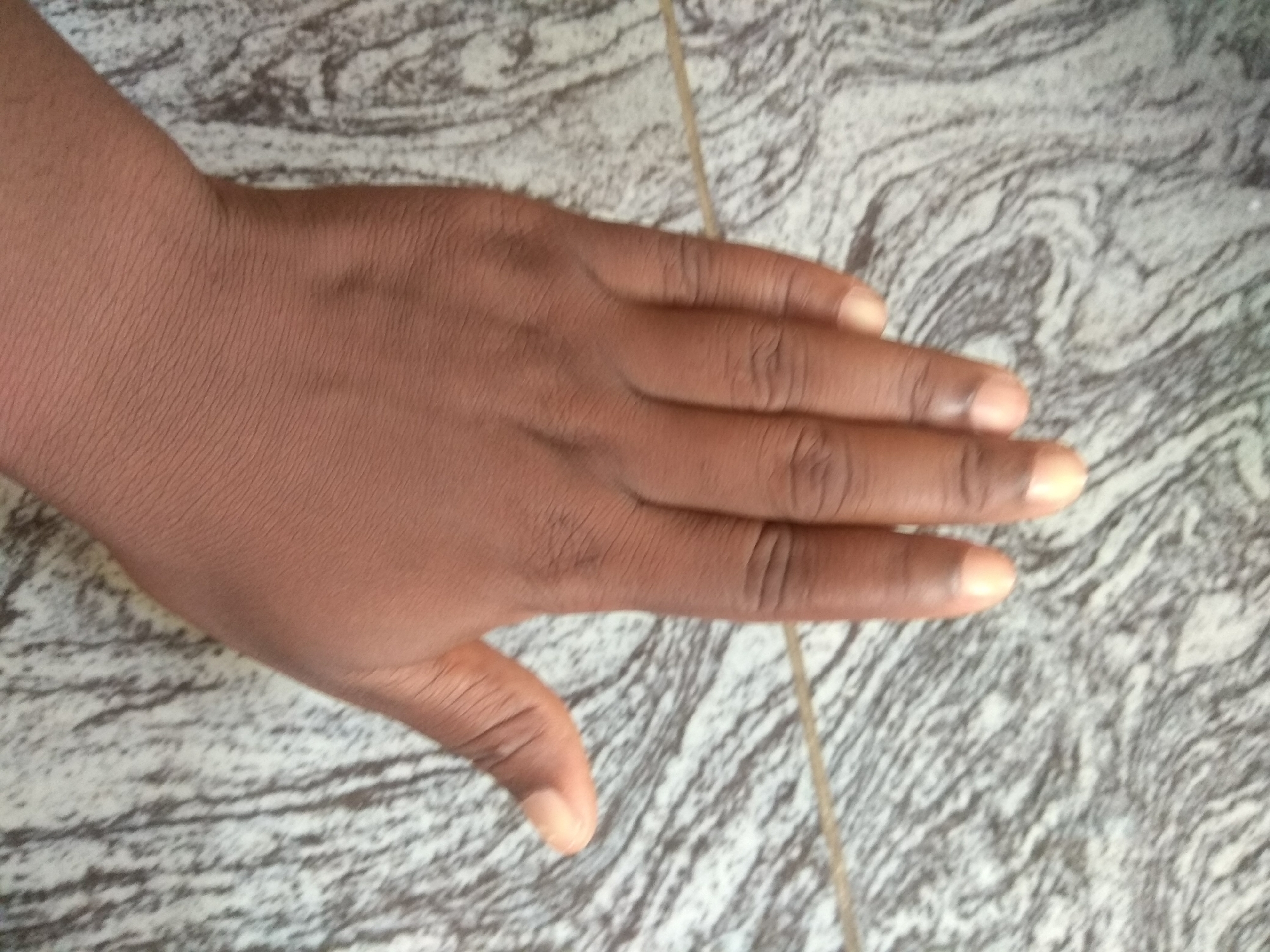 A close-up, overhead shot focuses on a dark-skinned person's hand resting on a patterned floor. The hand is positioned with the palm facing upwards and slightly towards the viewer, with the fingers gently curled. The skin on the back of the hand exhibits fine wrinkles and a light dusting of hair. The fingernails are visible, appearing clean and lightly colored. The floor beneath the hand features a swirling, marbled pattern of black, grey, and white. A thin, light-colored string or thread runs diagonally across the image, from the top right to the bottom left, partially obscured by the hand. The lighting is even, suggesting an indoor setting. The image was taken in Jalingo, Nigeria.