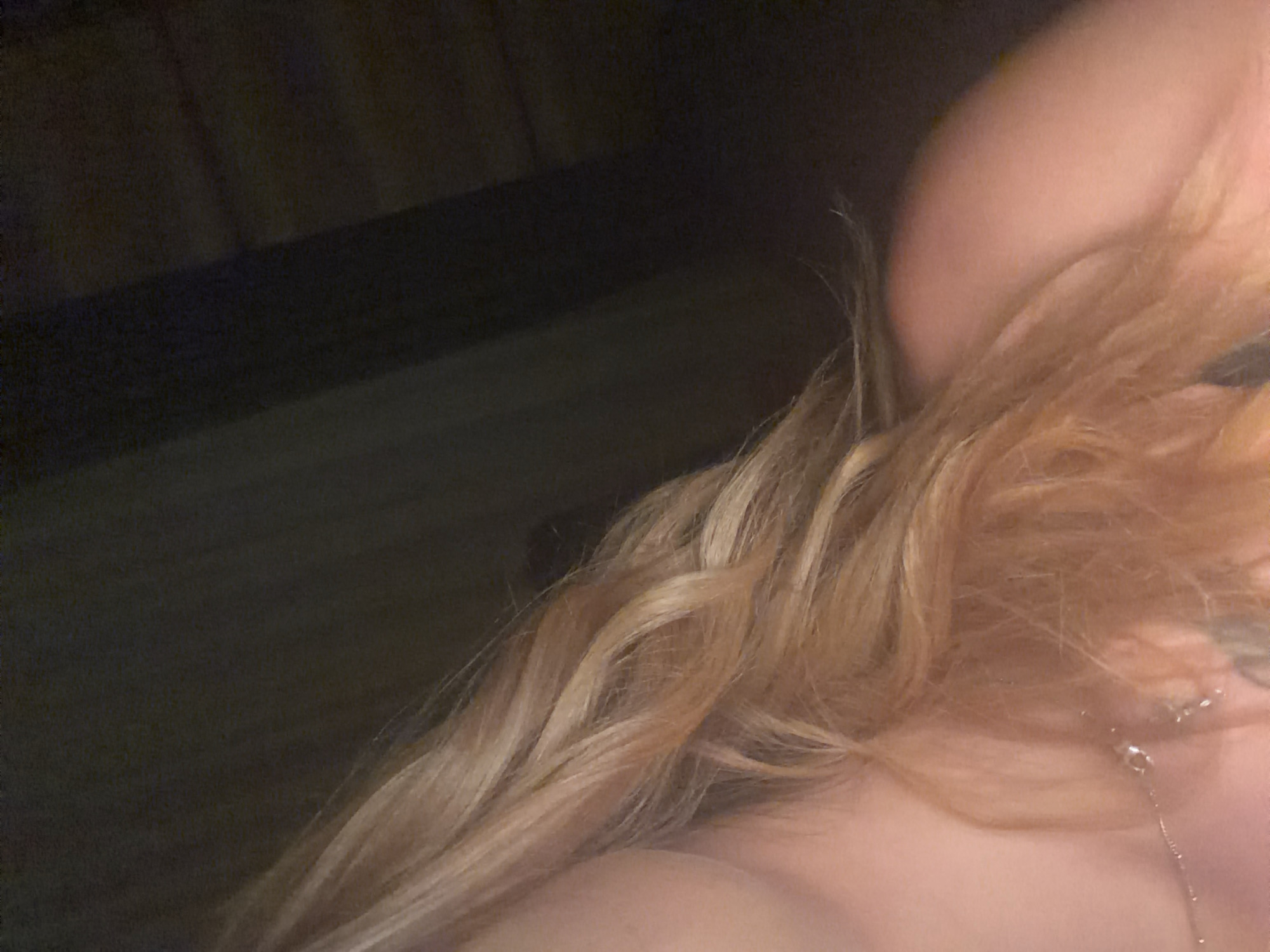 This is a close-up, slightly blurry image focusing on a person's shoulder and blonde hair. The background is dark and indistinct, suggesting an indoor setting with low lighting, possibly at night. The person's skin appears smooth, and their hair is styled in loose waves, with strands falling forward. A thin, silver necklace with a small charm is visible resting on the shoulder. The overall mood is intimate and personal, with no specific activity or event clearly depicted. There is no visible text in the image.