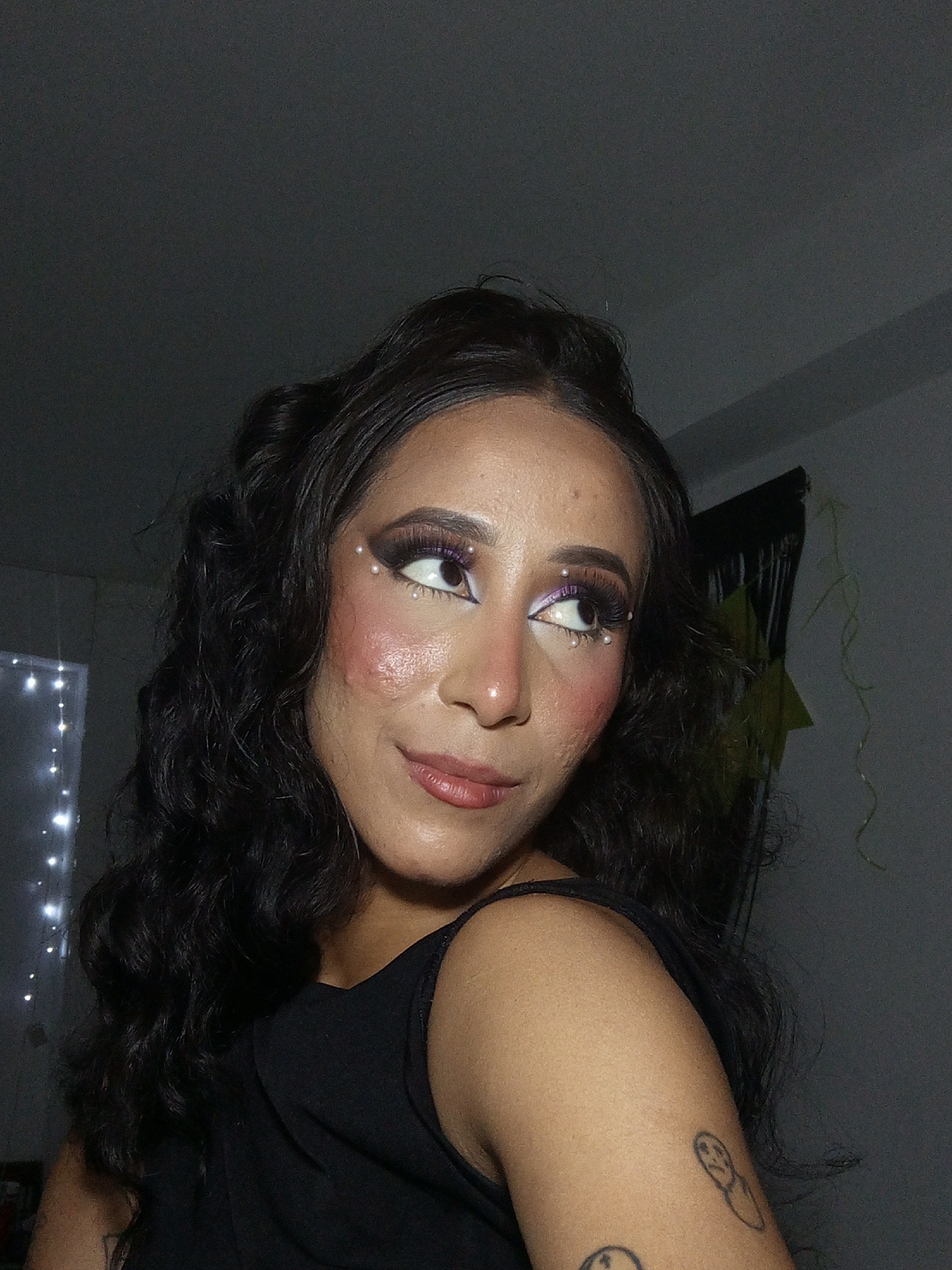 A woman with dark, wavy hair is facing away from the viewer but looking over her shoulder to the right. She has a black tank top on. Her makeup includes dramatic purple eyeshadow, false eyelashes, white pearls surrounding her eyes, and heavy blush. She has a tattoo on her upper arm. The background is a dimly lit room with what appears to be string lights and a green decoration hanging on the wall. It is not possible to determine the location in Porlamar, Venezuela from this image.