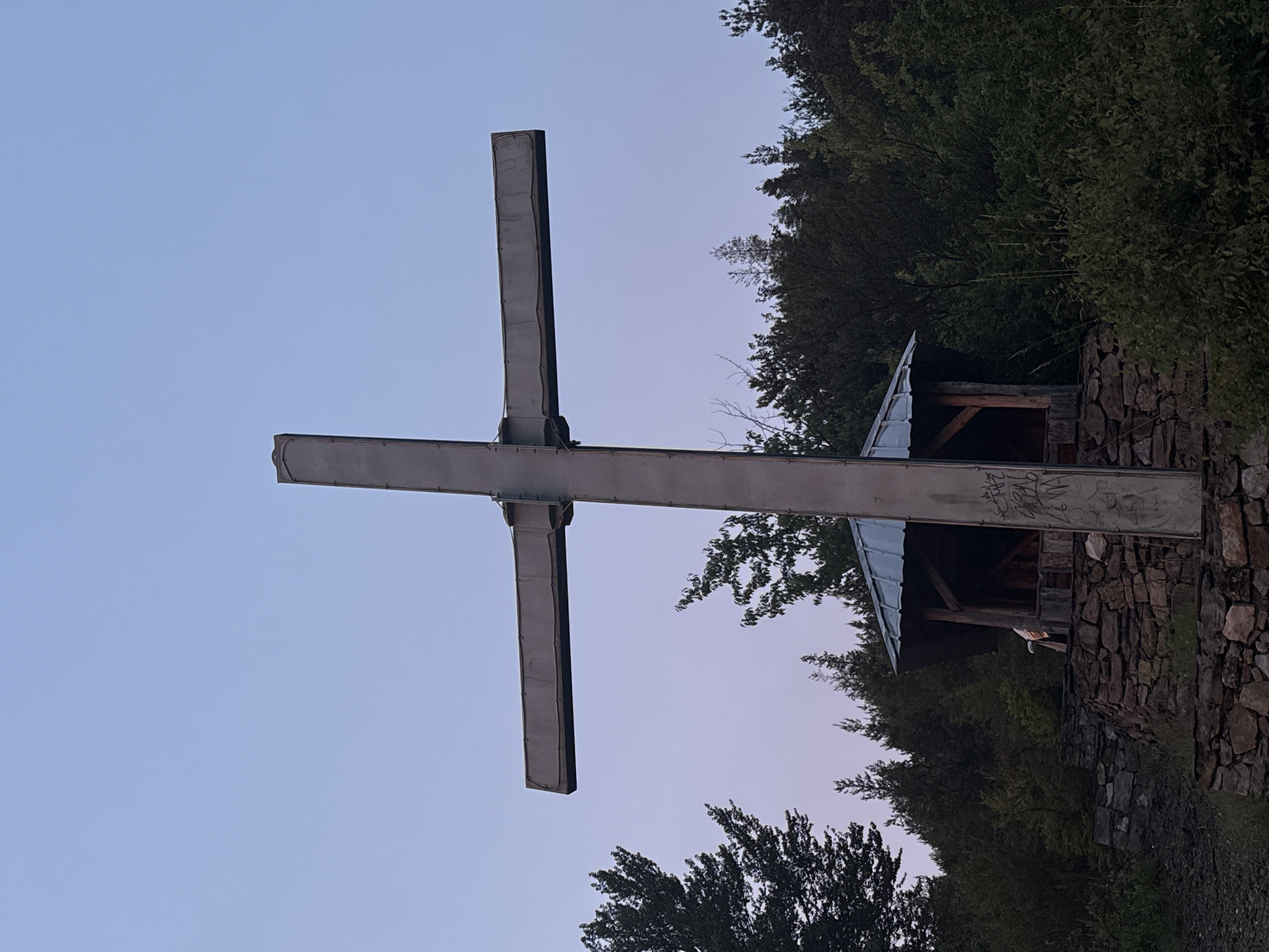 The image depicts a large, metal cross standing on a rocky, elevated area surrounded by trees. The cross is tall and appears to be made of industrial-style metal beams. At the base of the cross, there is a small, open-sided shelter with a metal roof, supported by wooden beams. The shelter is built on a stone foundation. The ground in front of the shelter is also covered in stones. A person can be partially seen inside the shelter. The sky above is a pale blue, transitioning to a soft purple hue near the horizon, suggesting either dawn or dusk. The surrounding environment is a dense forest, with various types of trees and foliage. The location is identified as being in the city of Strazha, Bulgaria.