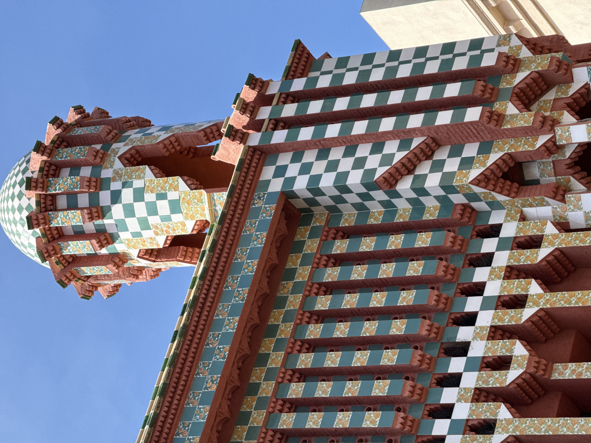 The image shows a close-up of the colorful facade of a building in Barcelona, Spain. The building features a prominent tower topped with a green and white checkered dome. The tower and the main body of the building are adorned with intricate patterns of turquoise, white, and orange ceramic tiles, arranged in a checkerboard and floral motif. Red brickwork and ornate architectural details, such as crenellations and stepped ledges, are also visible. The sky is a clear, bright blue. The building appears to be a historic or artistic structure, possibly a modernist architectural work, given its elaborate decoration and unique design. No people are present in the image.