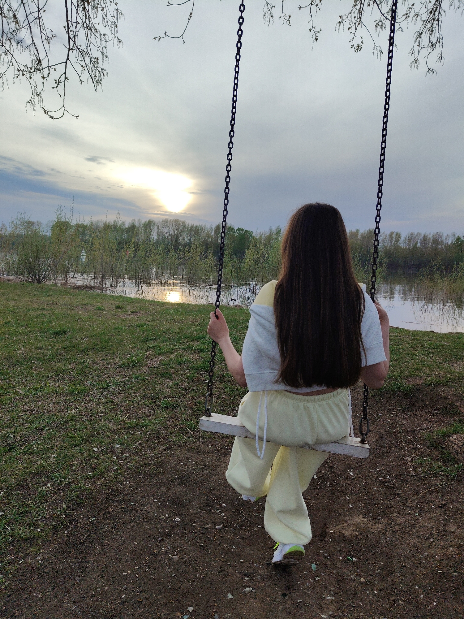 A woman with long, dark hair sits on a wooden swing, facing away from the viewer. The swing is suspended by two metal chains. She wears a light yellow sweat suit with a white t-shirt. She holds onto the swing chains with both hands. The swing is located in a grassy area with trees and water in the background. The sun is setting in the distance. The location is in the city of Unknown, Unknown.