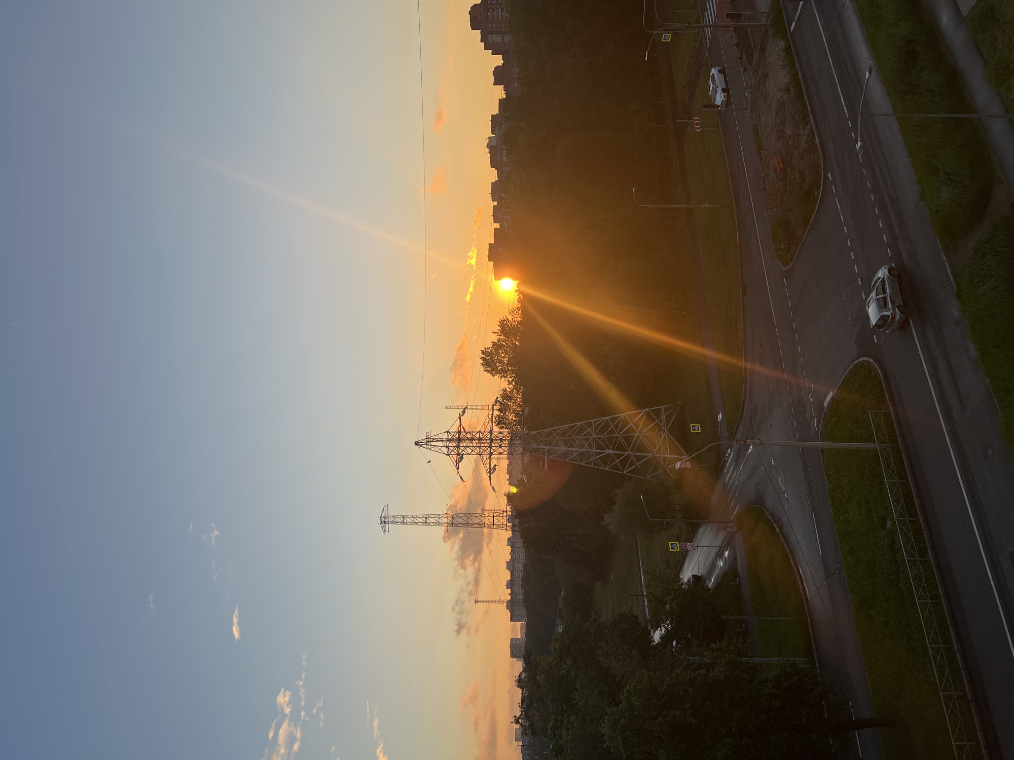 The image depicts a sunset over a city, likely Saint Petersburg, Russia. The sun, a bright orange orb, is low on the horizon, casting long rays of light across the sky and illuminating the scene. The sky is a gradient of blue and orange, with scattered clouds tinged with pink and yellow.
Dominating the foreground are several large metal electricity pylons, their skeletal structures silhouetted against the sunset. Trees and foliage fill the middle ground, creating a dense green band that separates the pylons from the urban landscape beyond. In the distance, a row of apartment buildings stretches across the horizon, their dark shapes contrasting with the bright sky.
A wet asphalt road with white lane markings curves through the scene, with two white cars visible. One car is parked on the side of the road, while the other is in motion. Streetlights and pedestrian crossing signs are also present, indicating an urban environment. The wetness of the road suggests recent rain or damp conditions.
The overall mood of the image is peaceful and serene, with the warm glow of the sunset creating a sense of calm. The combination of natural elements (sun, sky, trees) and man-made structures (pylons, buildings, roads) suggests an urban setting that is integrated with its natural surroundings. The time of day is clearly sunset due to the position and color of the sun and the long shadows. There is no visible text in the image.
