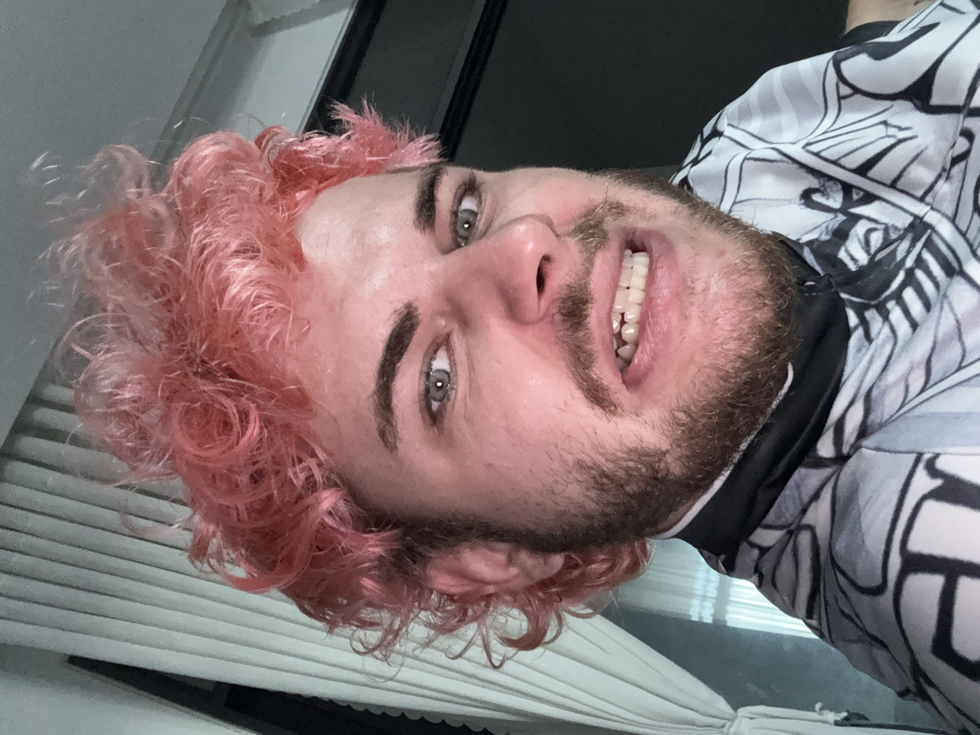 A close-up, low-angle selfie captures a person with fair skin, light eyes, a beard, and pink hair. The person is smiling, displaying their teeth. They are wearing a white shirt with a black geometric pattern. The background includes a partially visible window with closed blinds. There is no clear indication of the location being Đà Nẵng, Vietnam, or any other specific location.