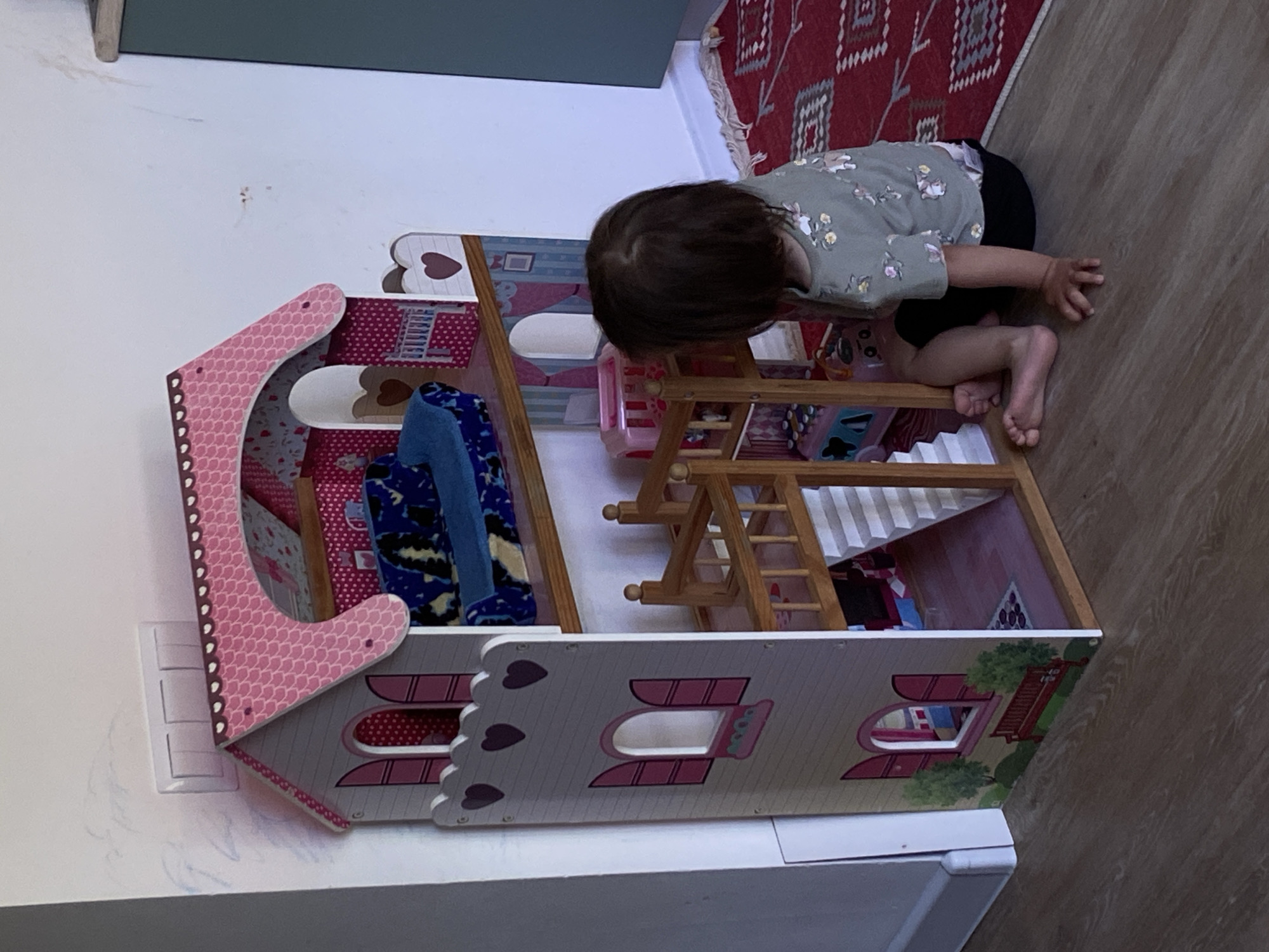 The image captures a young child engrossed in play with a dollhouse. The child, wearing a patterned shirt and dark shorts, is positioned on the floor, leaning over the dollhouse. Their head is close to the dollhouse, suggesting a focused interaction.
The dollhouse, primarily white with pink accents and heart-shaped details, is filled with various miniature items, including what appears to be furniture and accessories. The house is placed near a white wall, with an electrical outlet visible. A red rug with a geometric pattern is partially visible in the background.
The setting appears to be indoors, likely a home. Without additional landmarks or details, determining the precise location within Tyumen, Russia, is not possible.