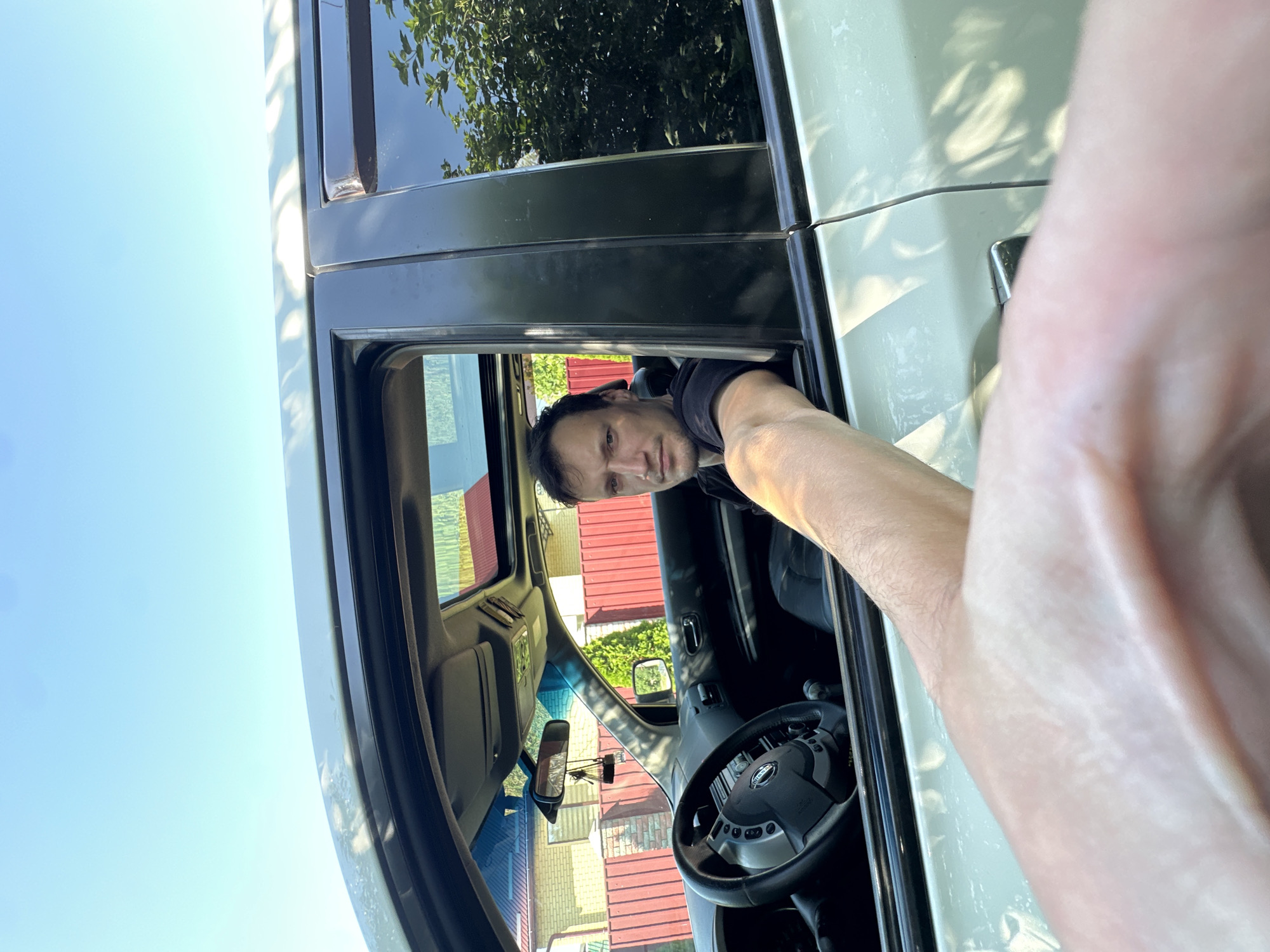 A man is sitting in the driver's seat of a car, with his left arm extended out of the open window, holding the camera. The car's sunroof is open, revealing a clear blue sky. The man is wearing a black t-shirt and looking directly at the camera. The car's interior includes a steering wheel and a rearview mirror. Outside the car, a brick house with a red fence is visible in the background, along with trees and foliage. The scene appears to be in a residential area during the daytime.