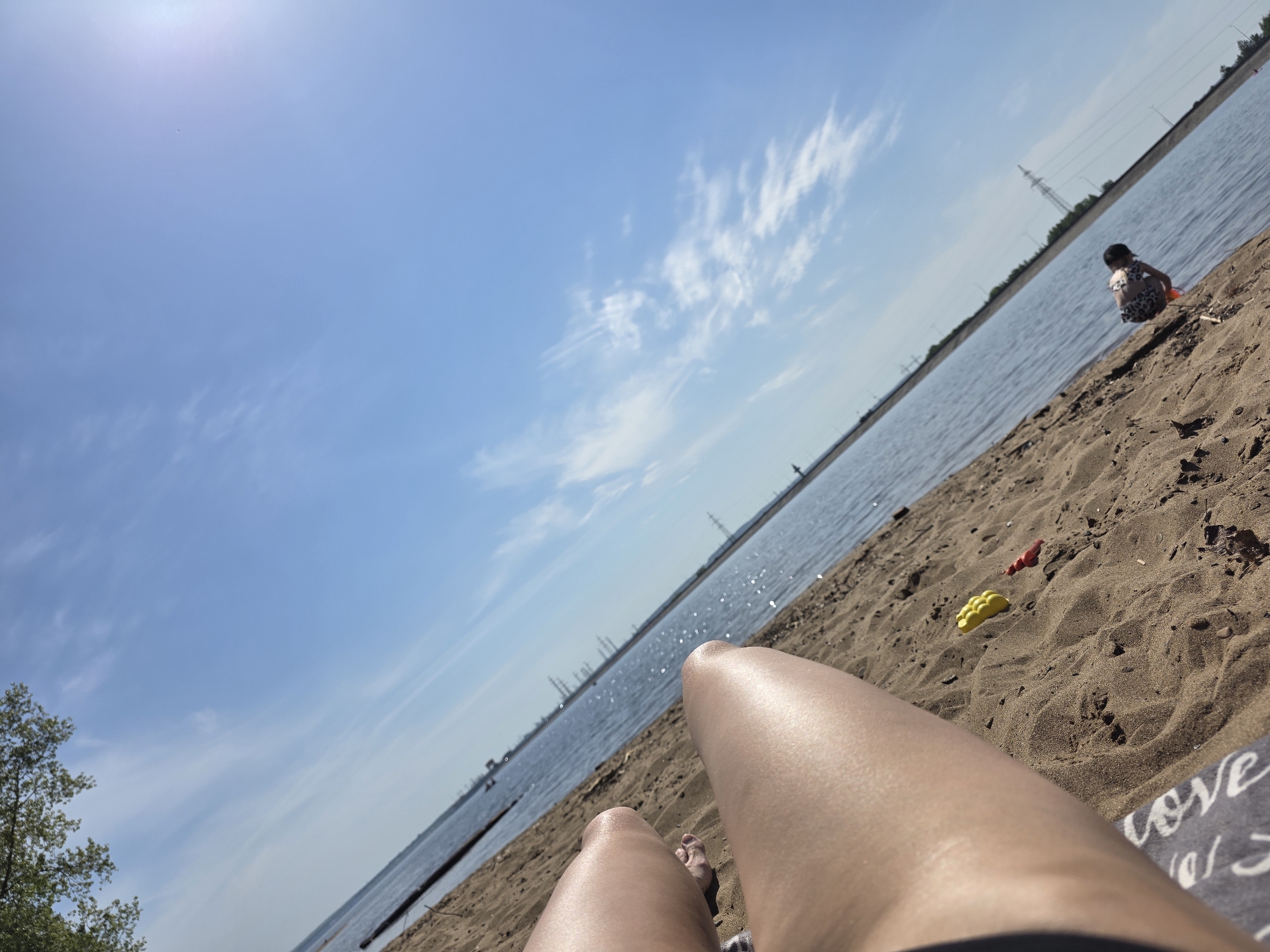 The image shows a sunny beach scene. A person is lying on the sand, their legs visible in the foreground. In the background, there is a body of water, likely a lake or river, with the opposite shore visible. There is a child playing near the water's edge. The shoreline is sandy with scattered debris. In the background, there are power lines visible, indicating an urban or industrial area nearby. The sky is blue with scattered clouds. The towel lying near the person's legs has the word "LOVE" printed on it.
While the prompt requests the location in the city of Noviy, Russia, there is no information in the image to confirm this. The location can't be determined from the image.