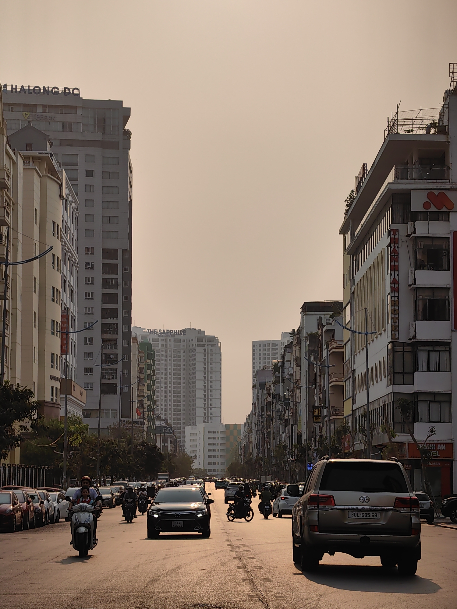 The image depicts a street scene in Hong Gai Ward, Vietnam, characterized by dense urban development. The view is down a wide avenue, with buildings lining both sides. On the left, a taller building with the inscription "HALONG DC" is visible, followed by other high-rise structures, including "THE SAPPHIRE" residences. On the right, buildings of varying heights are seen, some with commercial signage like "LANHEON" and "MSB ATM." The sky has a uniform, light hue. Traffic is present on the road, consisting of cars and scooters, with a Land Cruiser in the foreground with the license plate "30L-685.68" and a Toyota with the license plate "14A 526 24". There is a person on a scooter in the foreground of the image with a passenger riding in front of them.