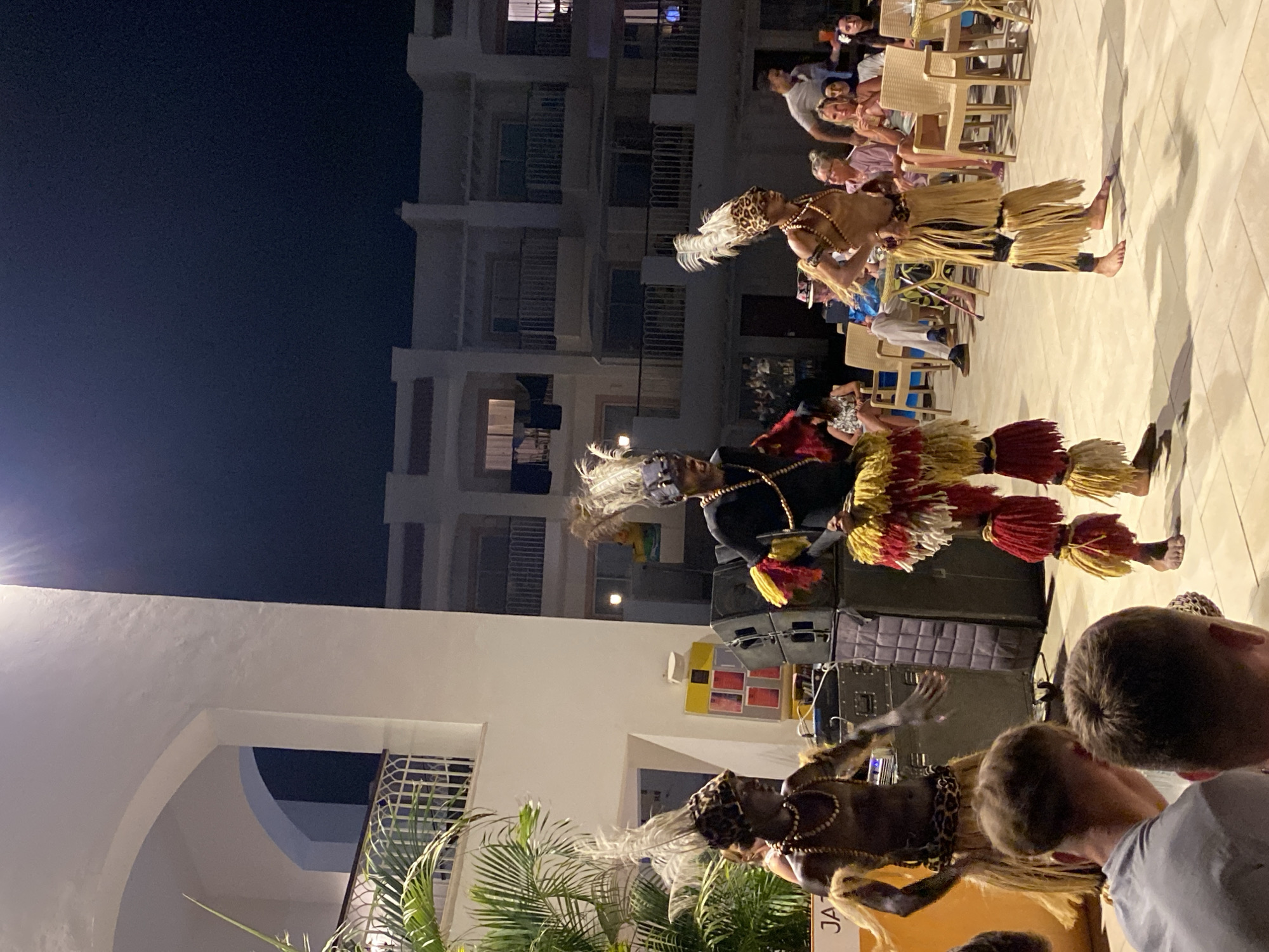 In Sharm El Sheikh, Egypt, an outdoor evening performance takes place in front of a multi-story white building with balconies. Three men in traditional attire, featuring feathered headdresses, beaded necklaces, and fringed skirts in red, yellow, and brown, are dancing. Two of the dancers are positioned in the foreground, while a third is slightly behind them. Their faces are dark, and their bodies are adorned with leopard print patterns and beads. Behind the dancers, a sound system with large speakers is set up. A banner with the partial word "JAZ" is visible on the left. Spectators are seated on chairs in the mid-ground, observing the performance. The lighting suggests it is nighttime, with some artificial lights illuminating the scene and the building. The ground is paved with light-colored tiles.
