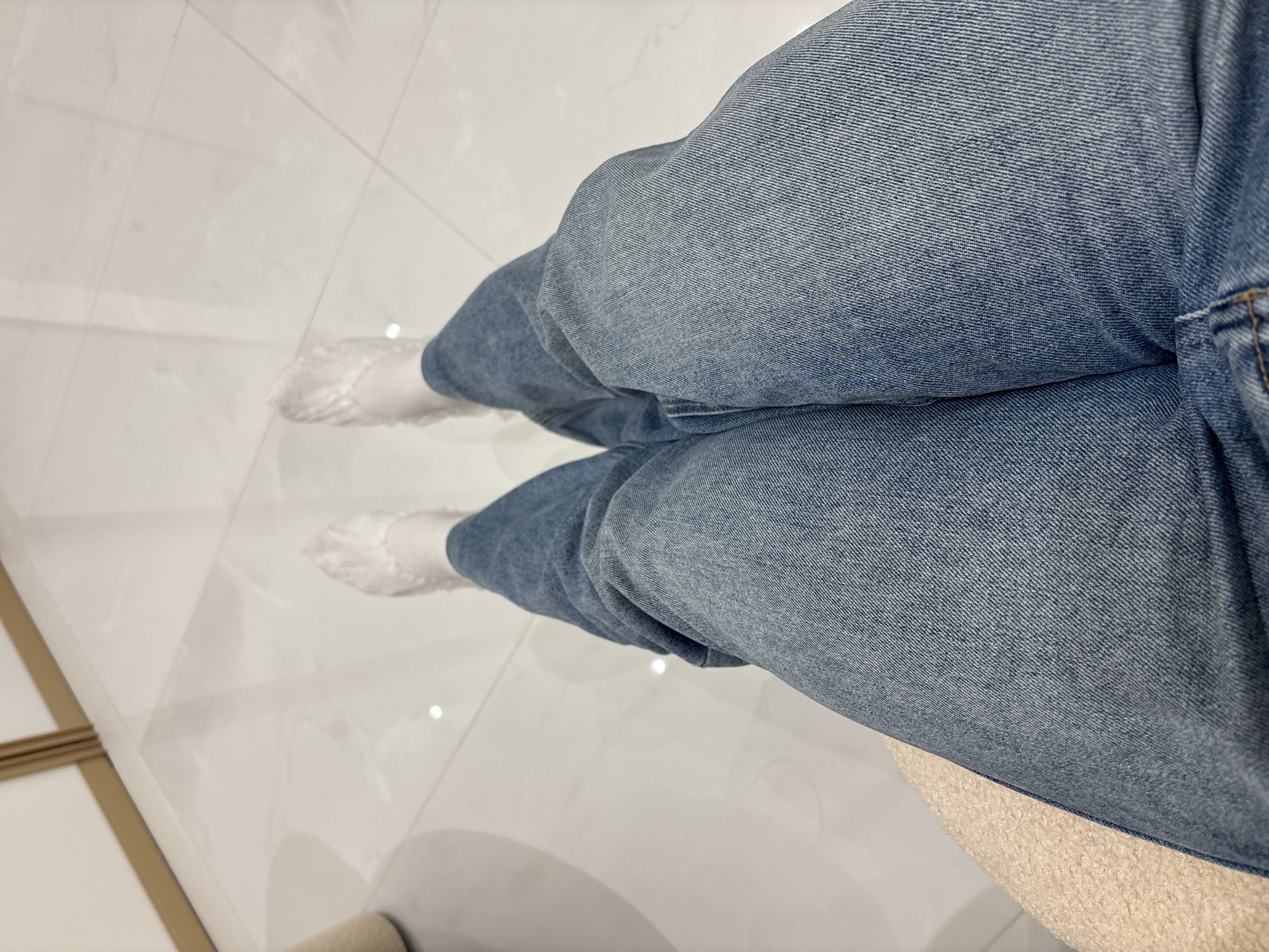 The image shows a person sitting, wearing light blue jeans and white disposable shoe covers, on a white tiled floor. The location is indicated to be Perm, Russia, but there are no details in the image to confirm this. The person's feet are visible in the shoe covers, and the lower part of their legs are covered in jeans. The surface beneath them is partially visible and appears to be a beige, textured chair.